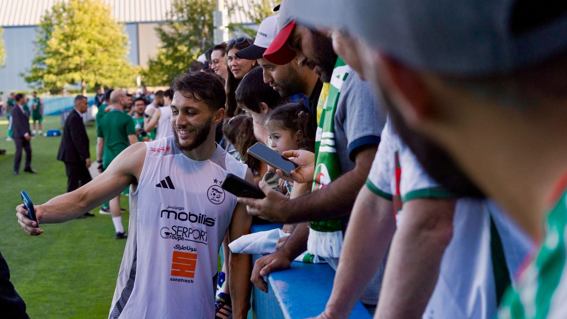 Smiling male athlete in a white sleeveless jersey takes a selfie with fans along a blue barrier; a young girl watches as others hold up phones at a sunny outdoor soccer field.