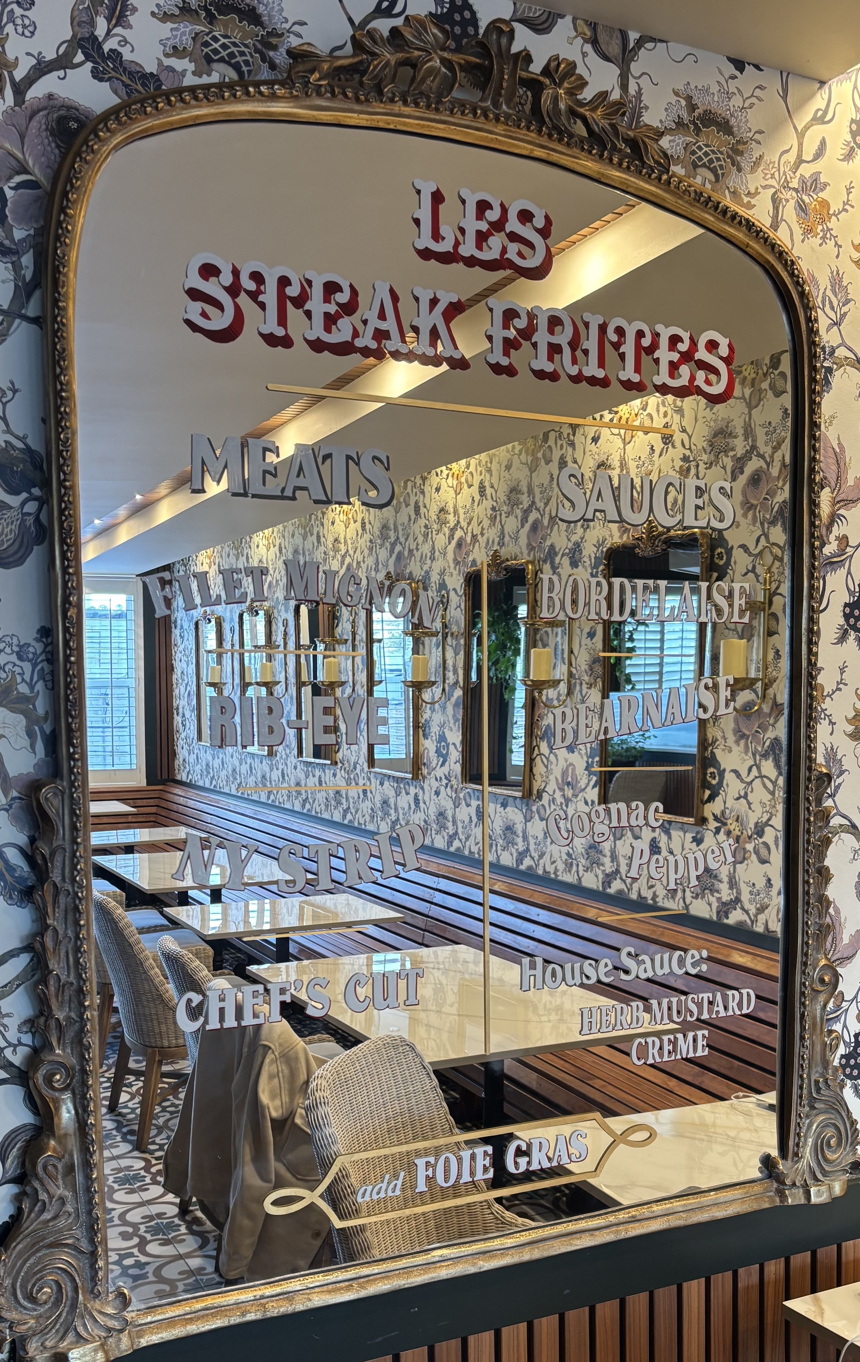 Ornate mirror with gold frame in a restaurant listing meats and sauces for "Les Steak Frites" on its surface, reflecting floral wallpaper, wooden bench seating, and tables inside.