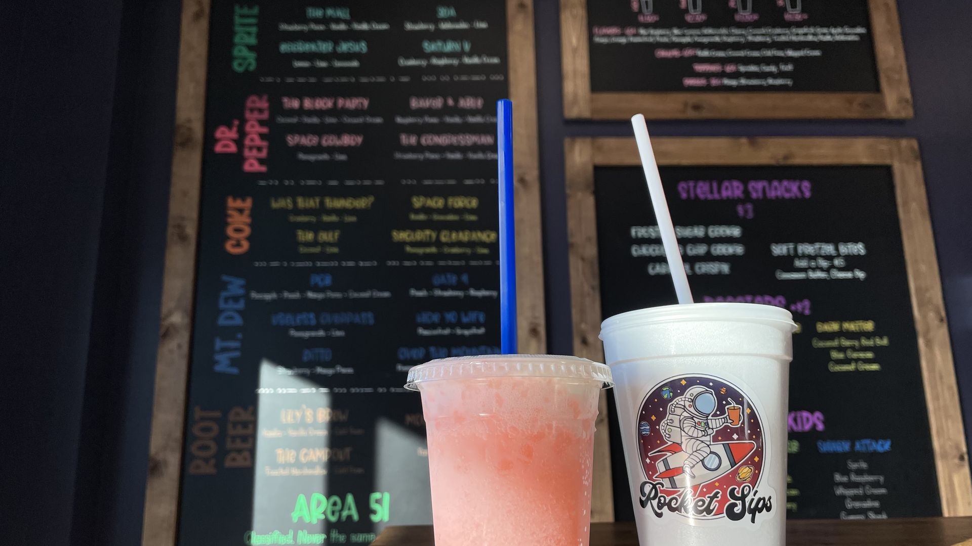 Two drinks on a wooden table in front of a menu board: a pink slushy with a blue straw in a clear cup and a white cup with an astronaut logo and white straw from Rocket Sips.