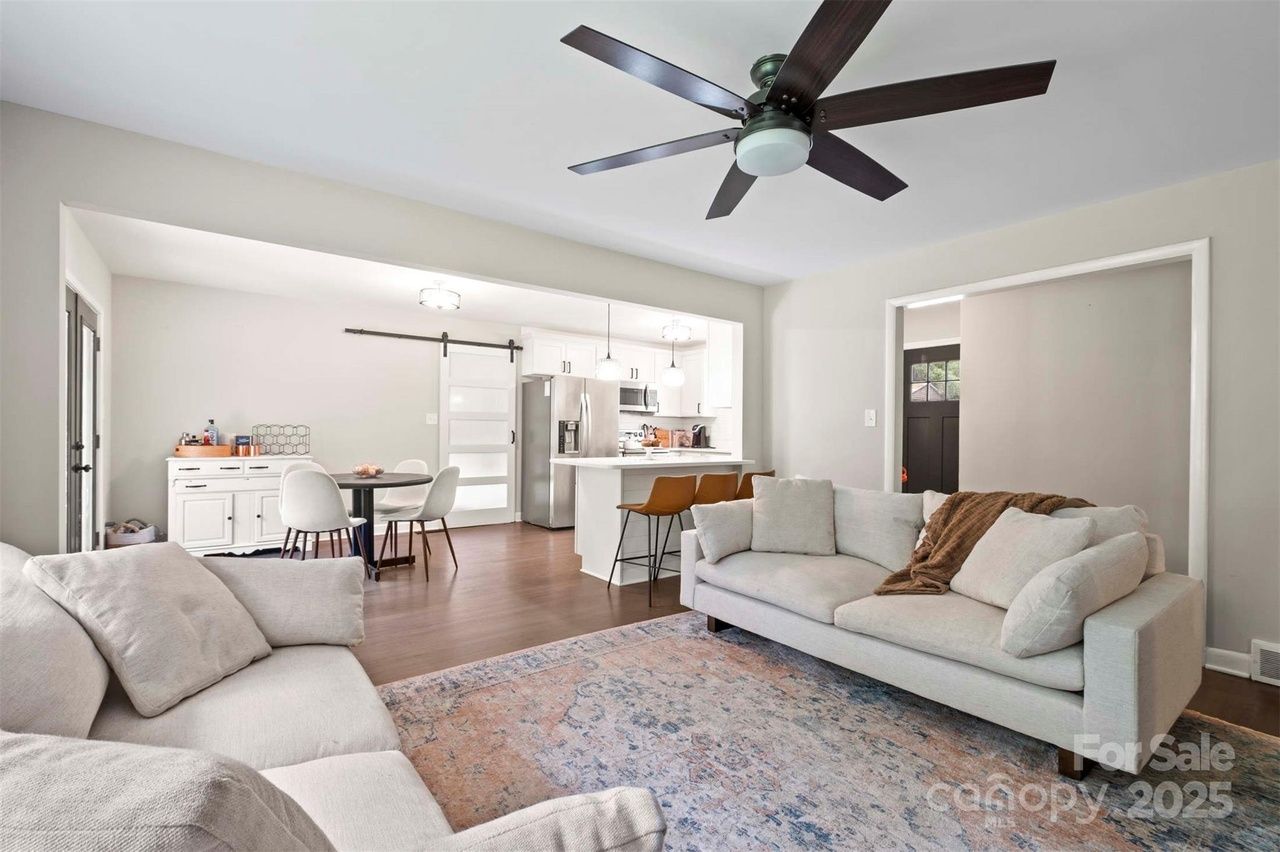 Bright open living room with beige sofas, a patterned rug, dark wood floor, ceiling fan, white kitchen with bar stools, and a small dining area with white chairs.