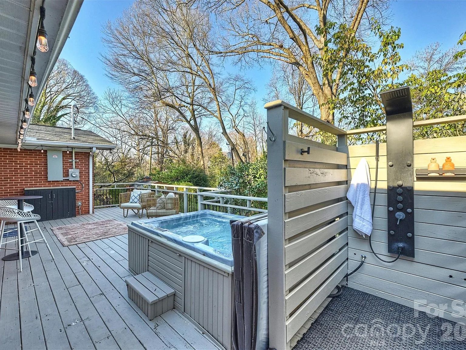 Outdoor wooden deck with a hot tub, two beige cushioned chairs, hanging string lights, a black outdoor shower, and a red brick house wall, surrounded by trees under a clear sky.