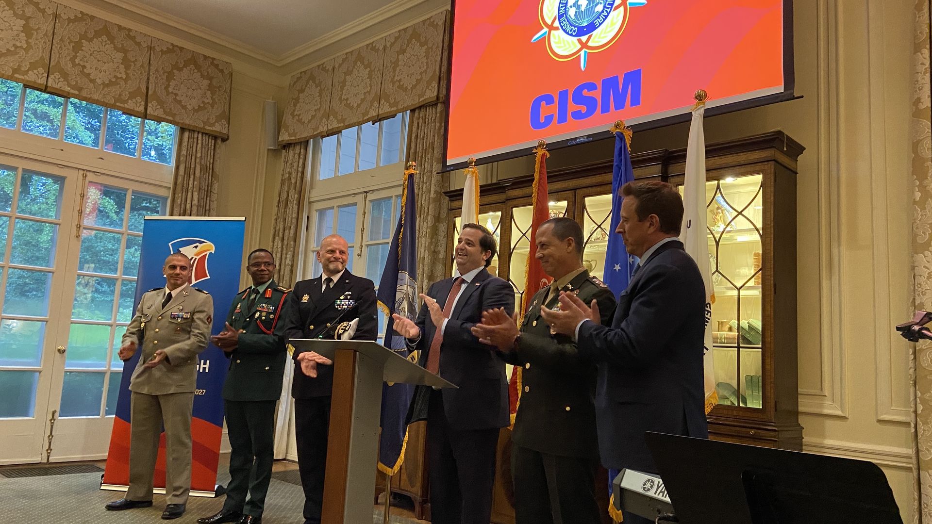 Six men in military and formal attire stand and clap in a decorated room with tall windows, flags, and a red screen displaying a CISM logo above them.