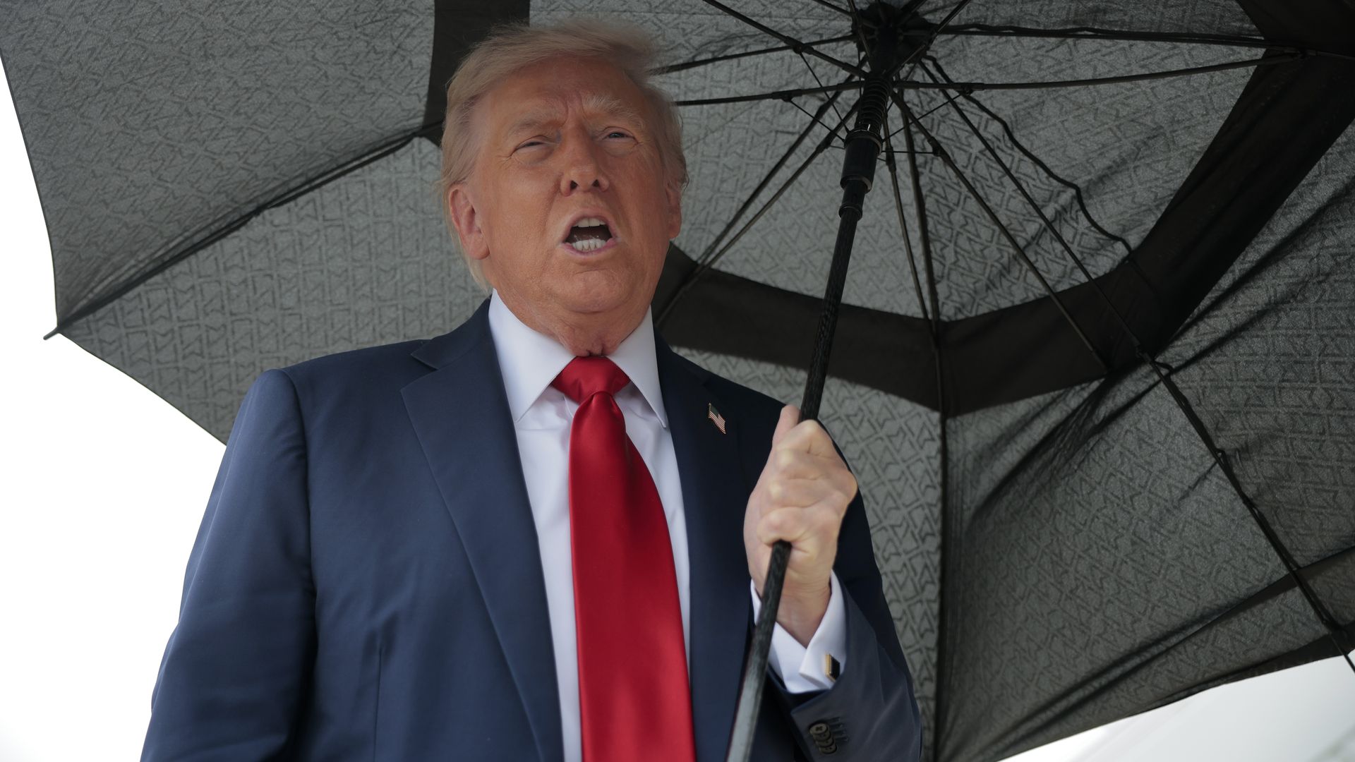President Trump, wearing a navy jacket with a US flag pin, white shirt and red tie, carries a black umbrella as he speaks to the press before boarding Air Force One for a trip to the Middle East on October 12, 2025 at Joint Base Andrews, Maryland. 