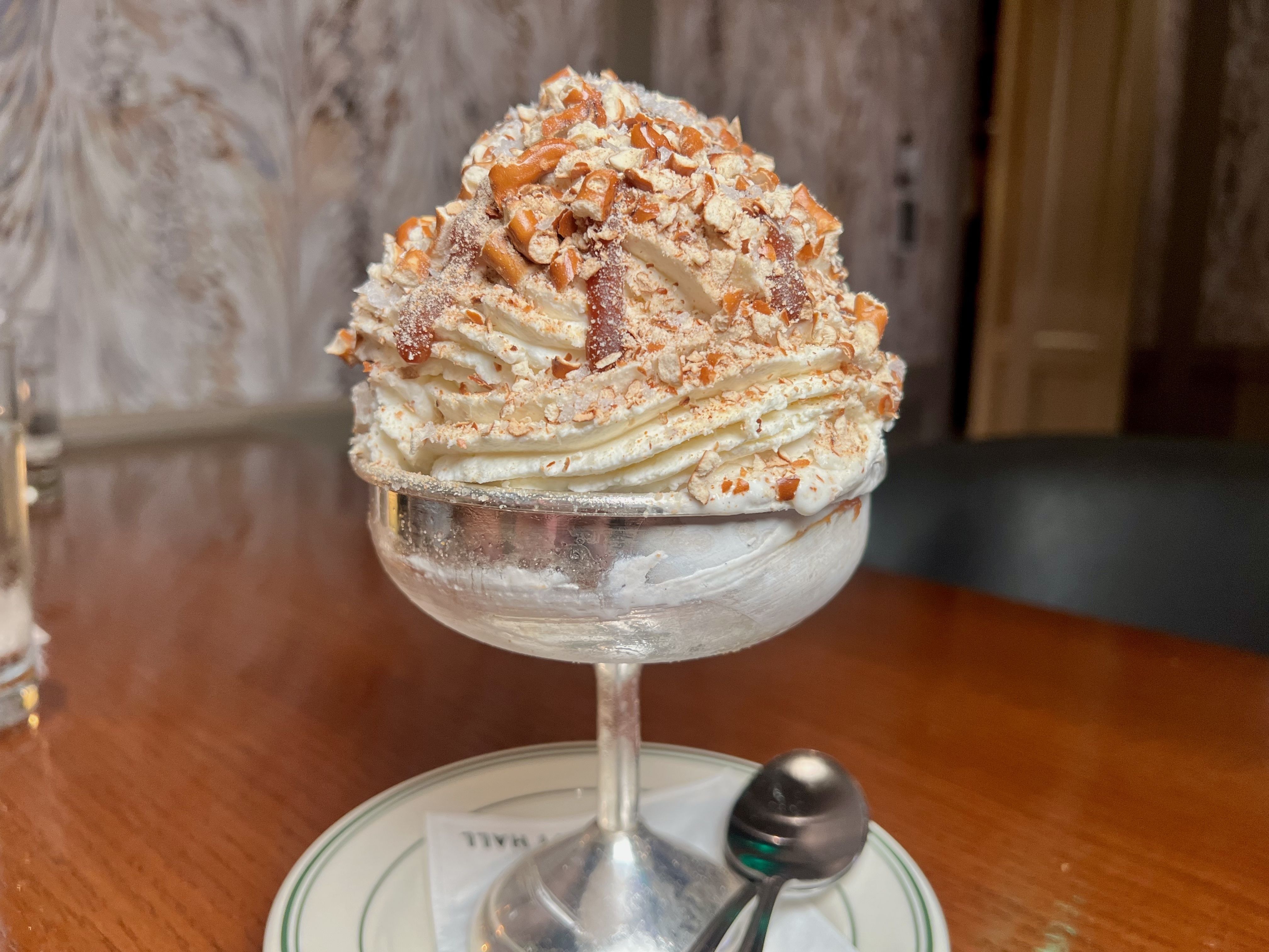 Whipped cream-topped dessert in a glass coupe, dusted with crushed caramelized toffee bits, resting on a white saucer with a spoon on a wooden table.