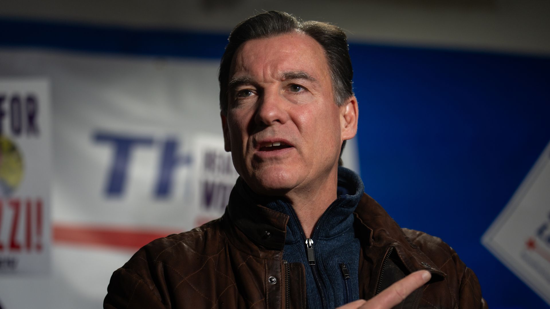Rep. Tom Suozzi, wearing a brown jacket over a blue fleece, standing in front of campaign signs and a blue backdrop.