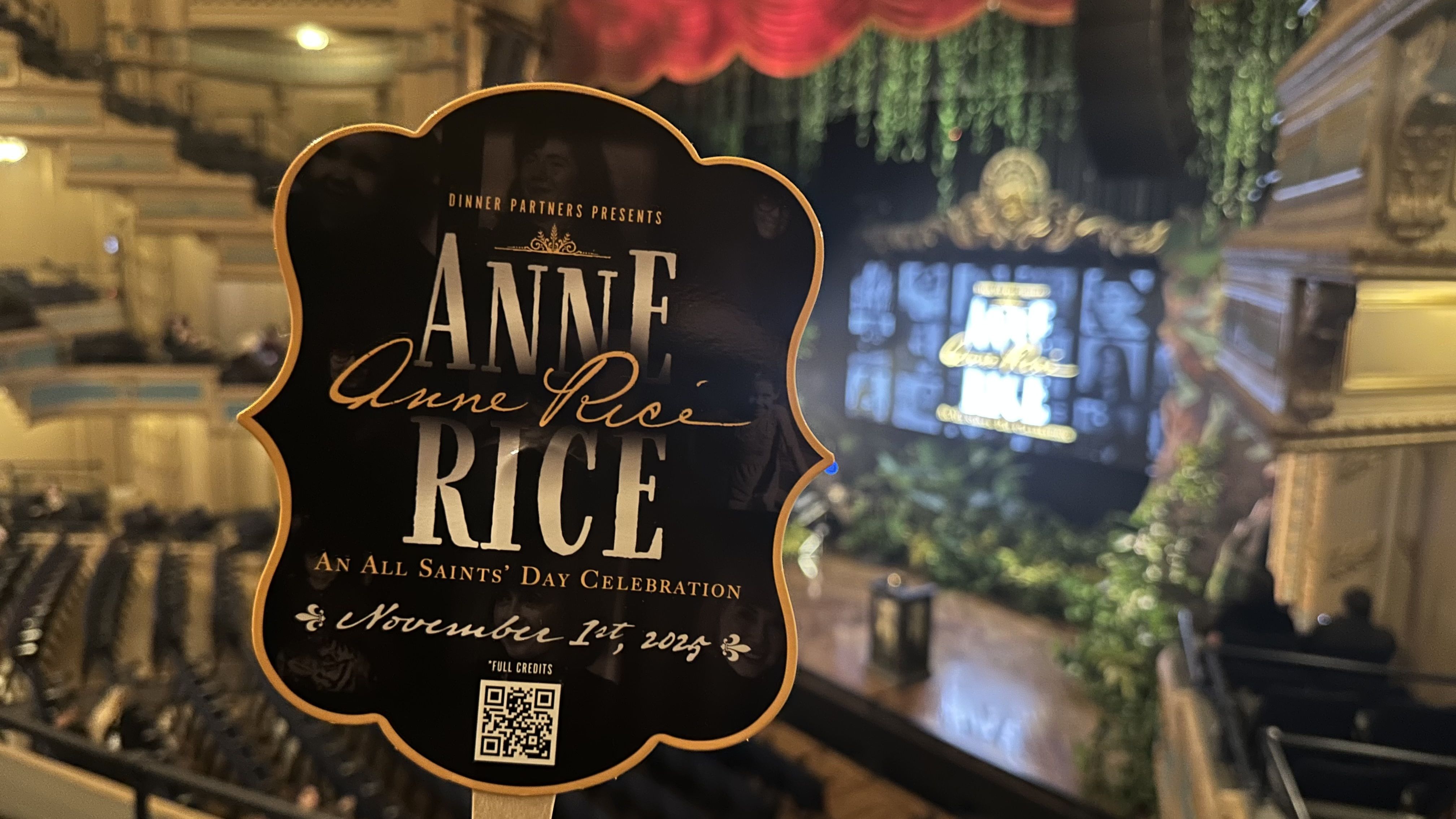 Sign in a theater announcing "Anne Rice: An All Saints' Day Celebration" on November 1st, 2023, with an ornate stage and audience seating in the background.