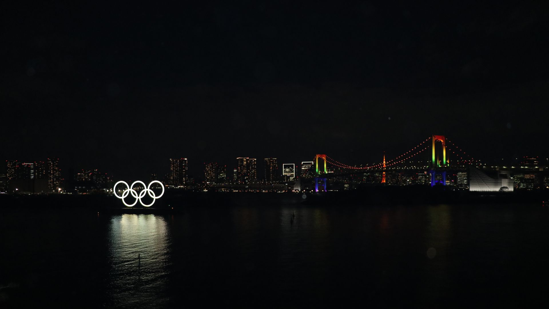 Picture of the olympic flag next to a bridge lit with rainbow lights