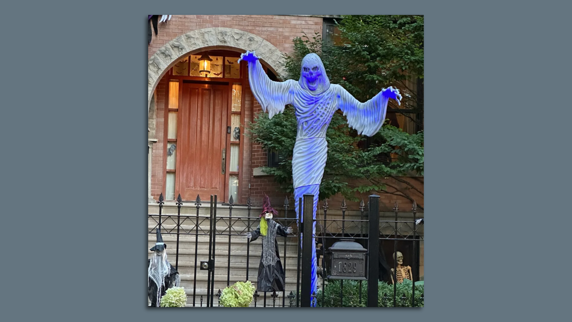 The front door of a house with doll witches hanging from the front gate and a spooky skeleton looming over them.