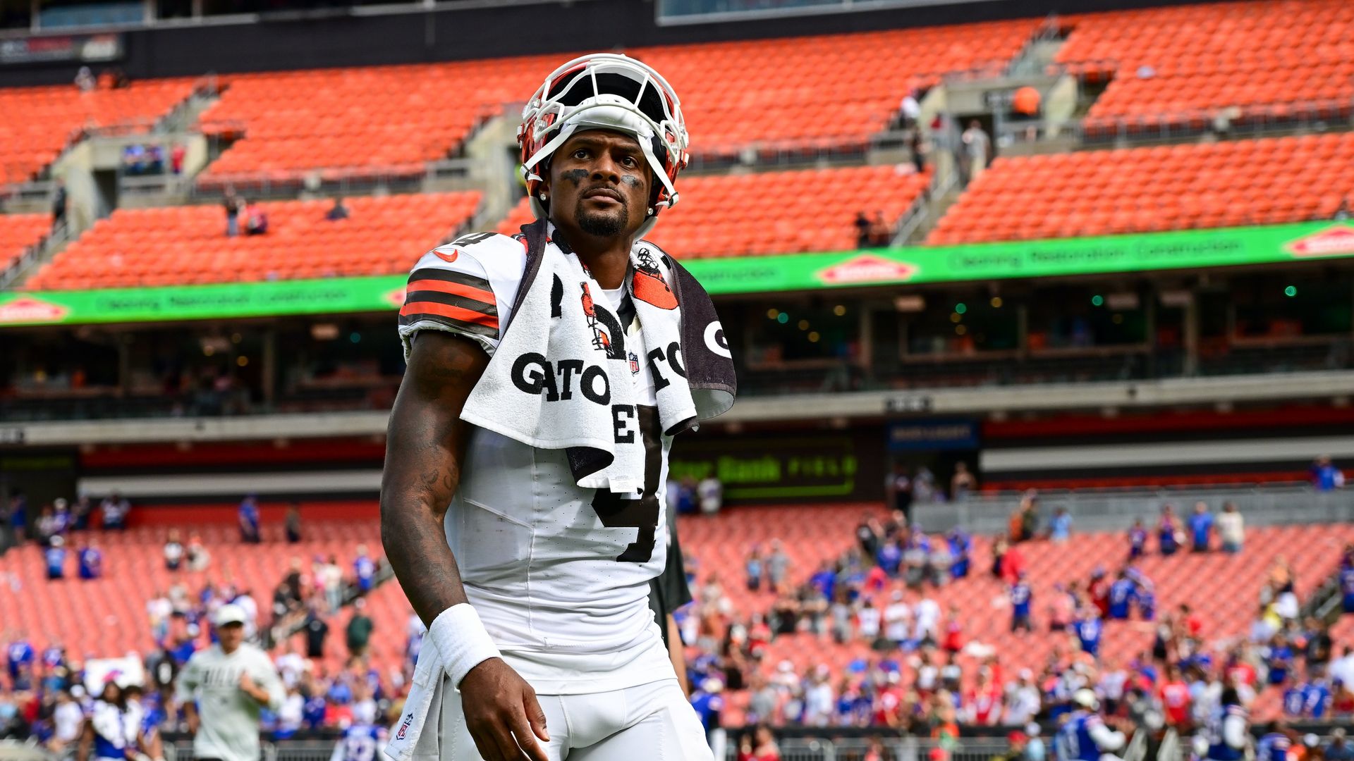 African American football player in a white uniform with orange and black stripes, helmet on, towel draped over shoulders, stands on a football field with orange stadium seats in the background.