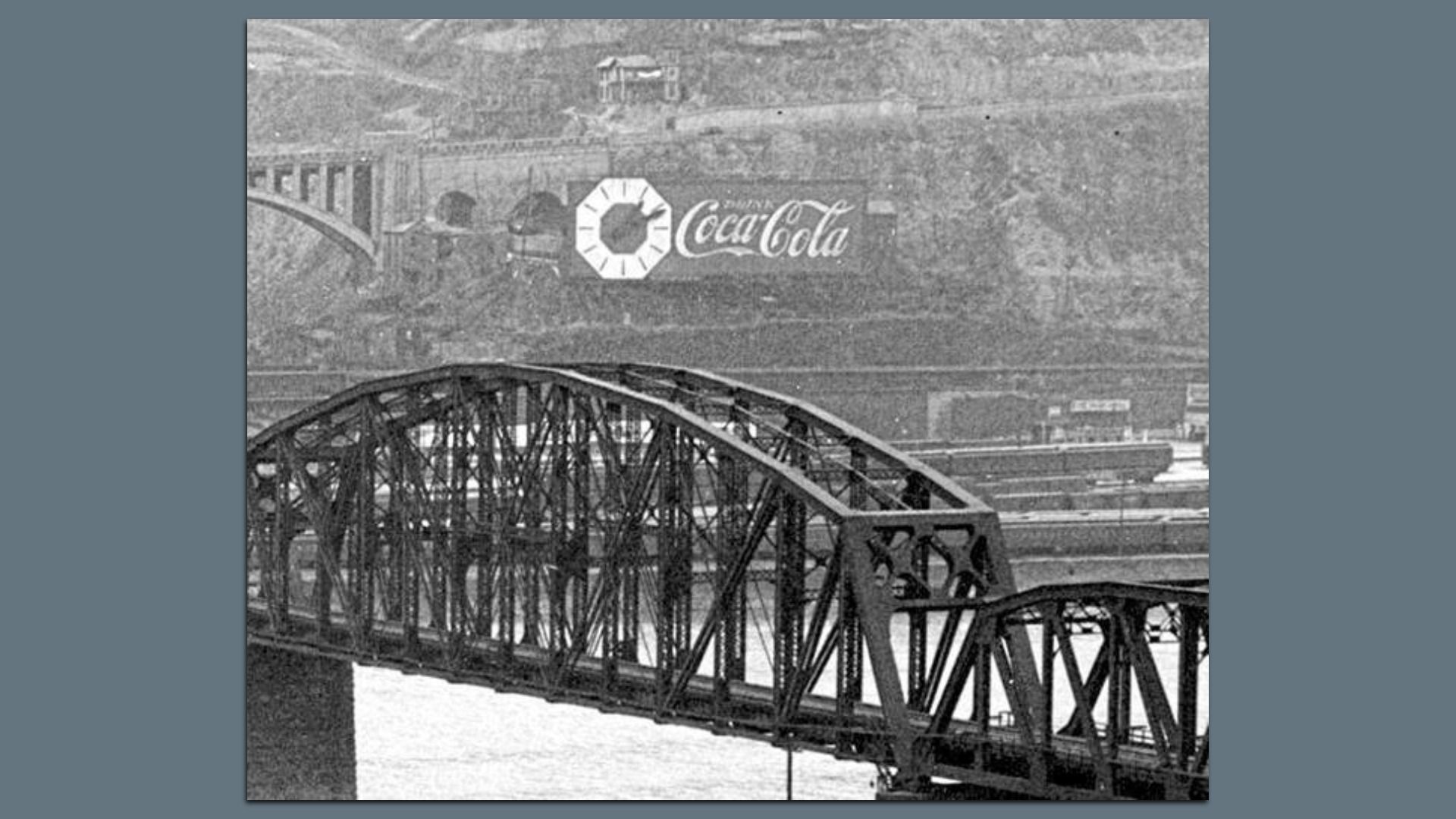 Black and white photo of a metal truss bridge over water with a hillside in the background showing a large Coca-Cola sign featuring a clock.