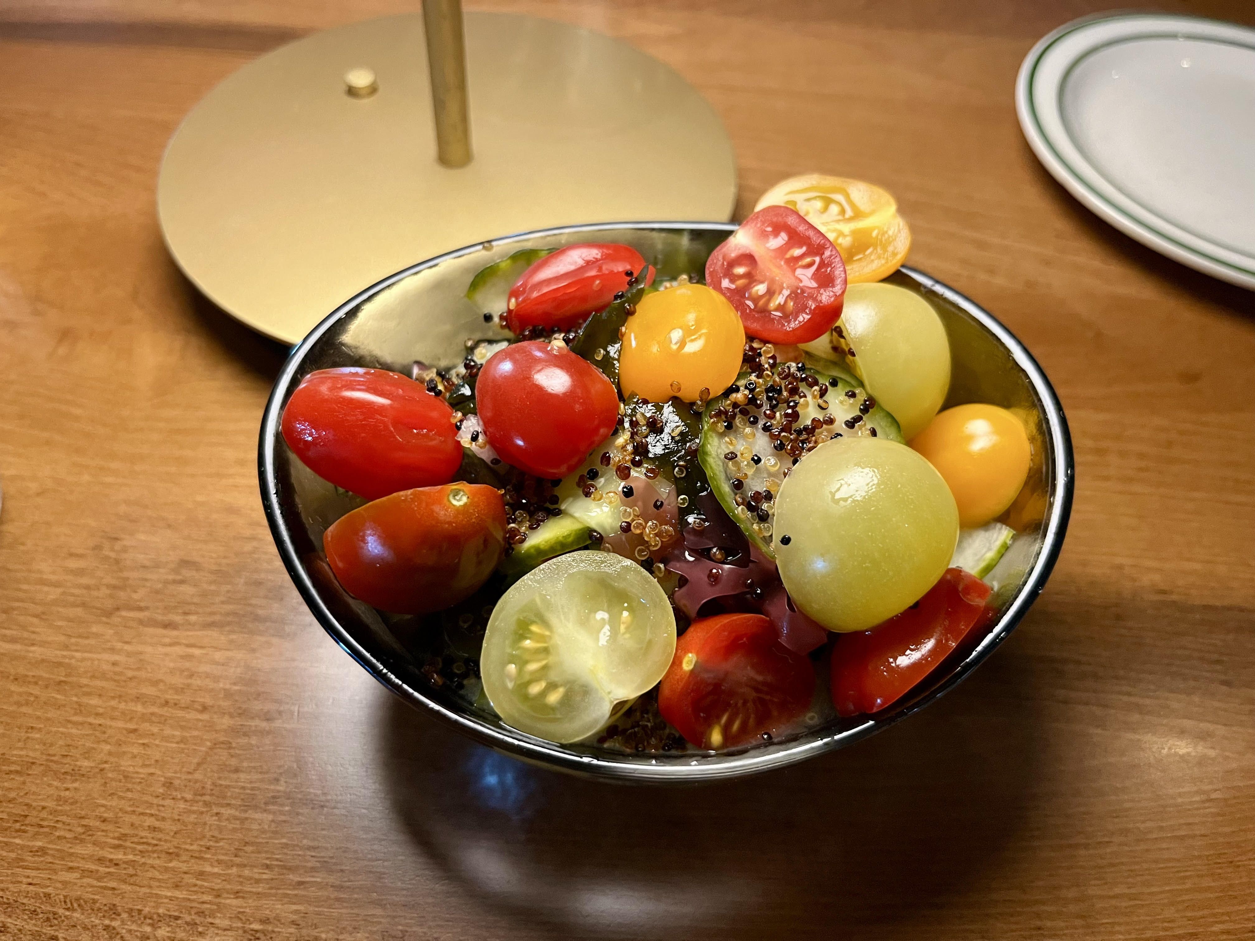 A silver bowl holds tomatoes, cucumbers, seeds and more on a wooden table under the light of a brass lamp.