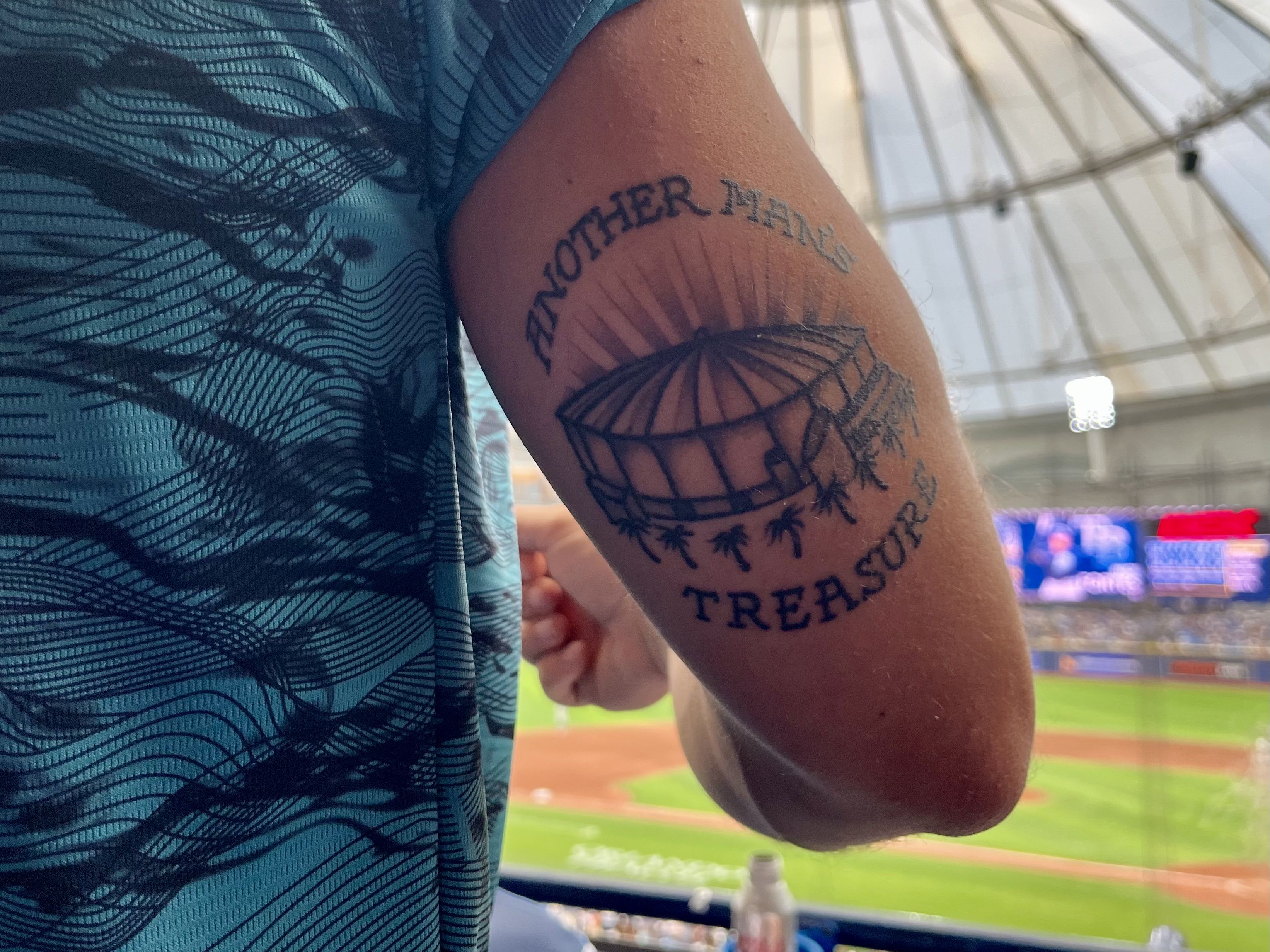 Tattoo on upper arm depicting a domed stadium with palm trees around its base; curved text above reads "ANOTHER MANS" and below "TREASURE".