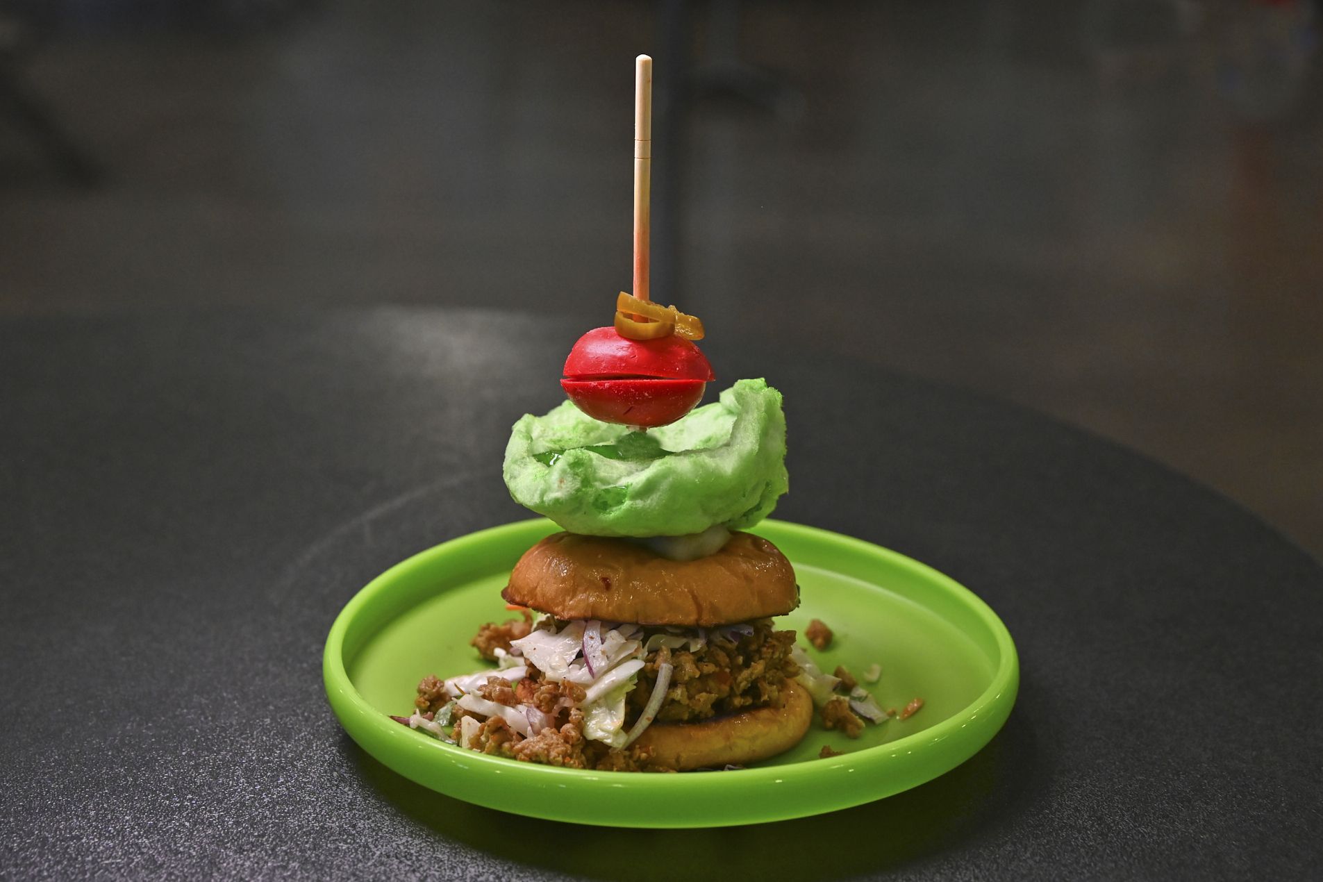 Stacked snack on a green plate: bottom bun with chopped meat and onions, a green ice-cream scoop, a red ball on a skewer with a yellow ring, set against a dark background.