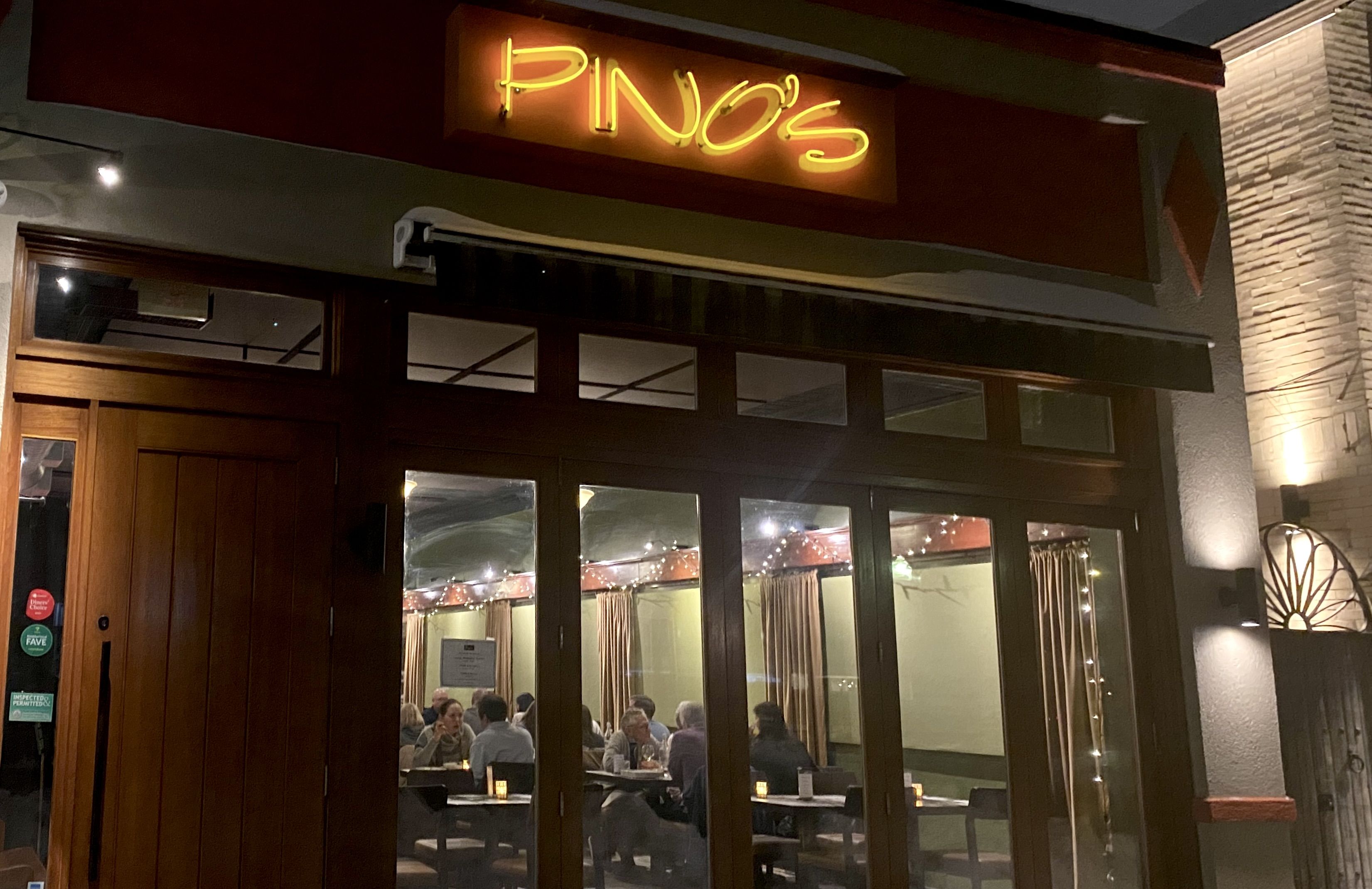 Night view of a warmly lit restaurant named PINO's with wooden framed glass doors and windows, showing diners inside and string lights hanging on beige curtains.