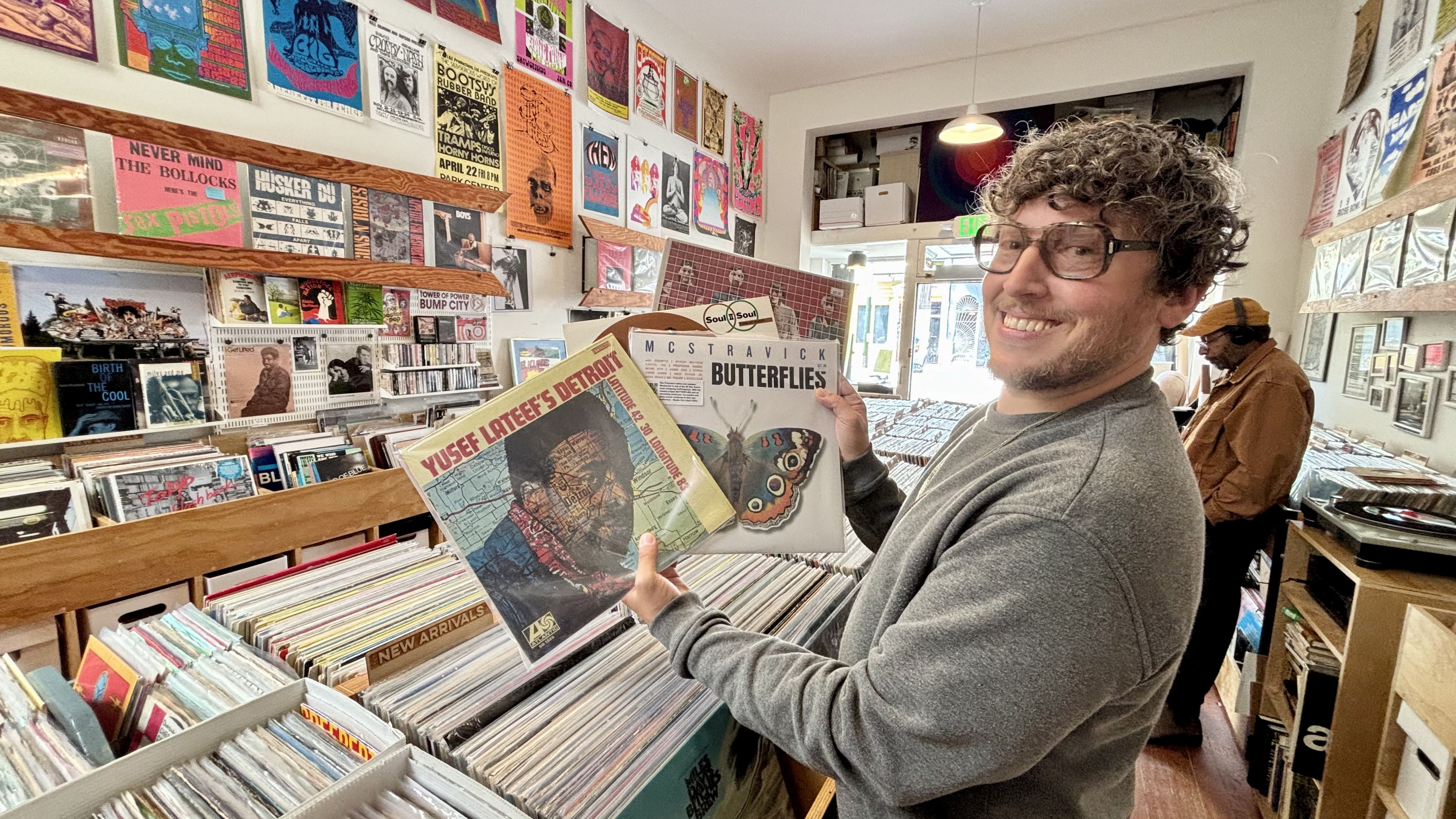 Man with glasses and curly hair smiles holding two vinyl records in a bright record store filled with colorful posters and rows of albums, another person browsing in the background.