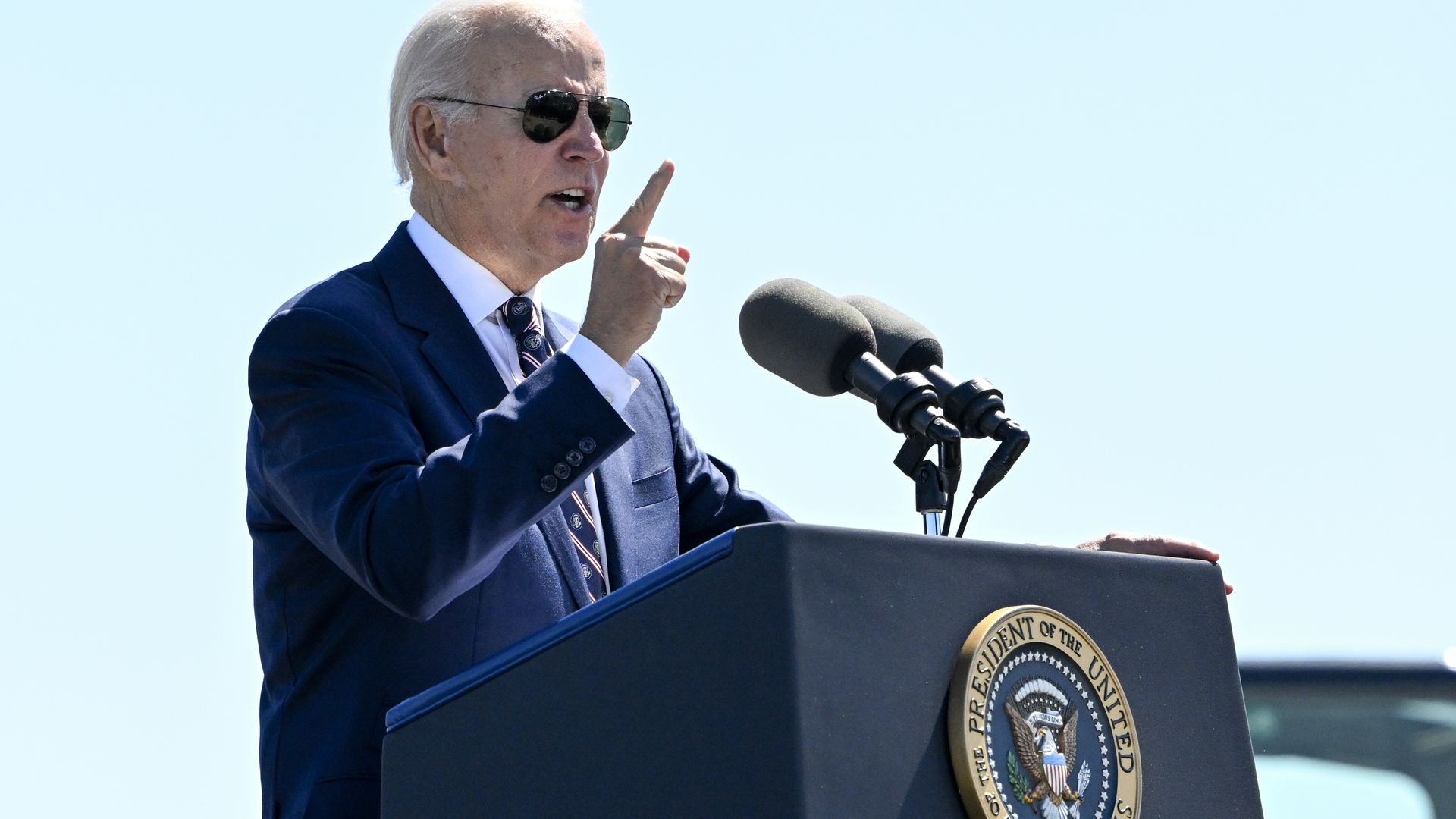 President Joe Biden raises his hand while speaking at a lectern. 