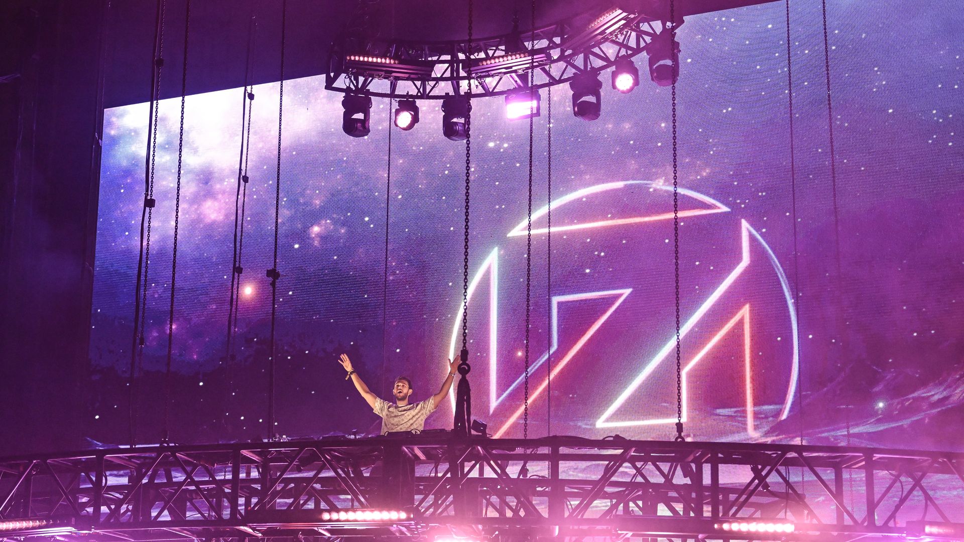 DJ performing on an elevated stage with bright pink and purple lights, a large circular neon logo in the background, and a starry cosmos-themed screen behind him.
