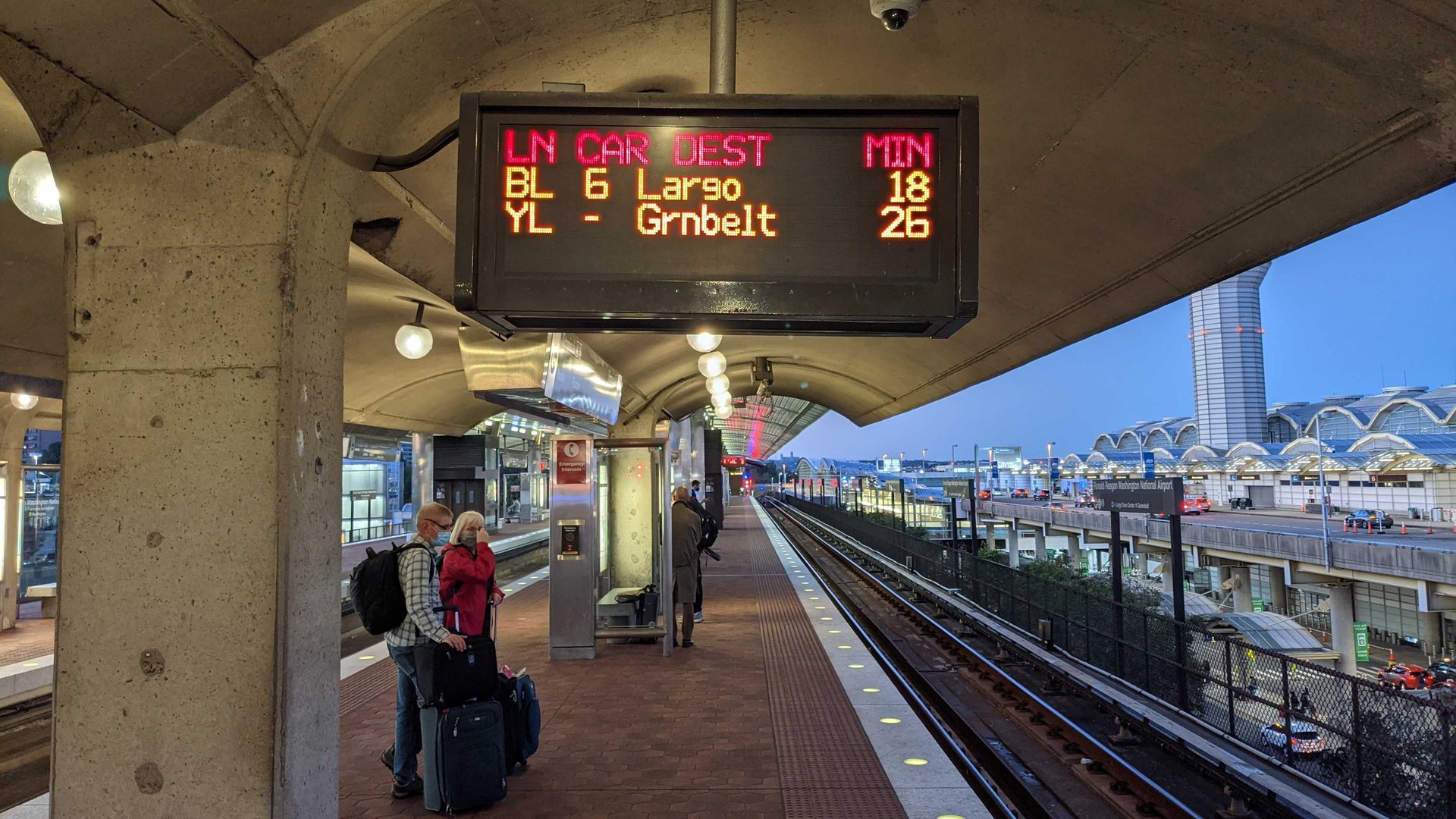 A Metro platform with long wait times. People stand with their luggage waiting for the train.