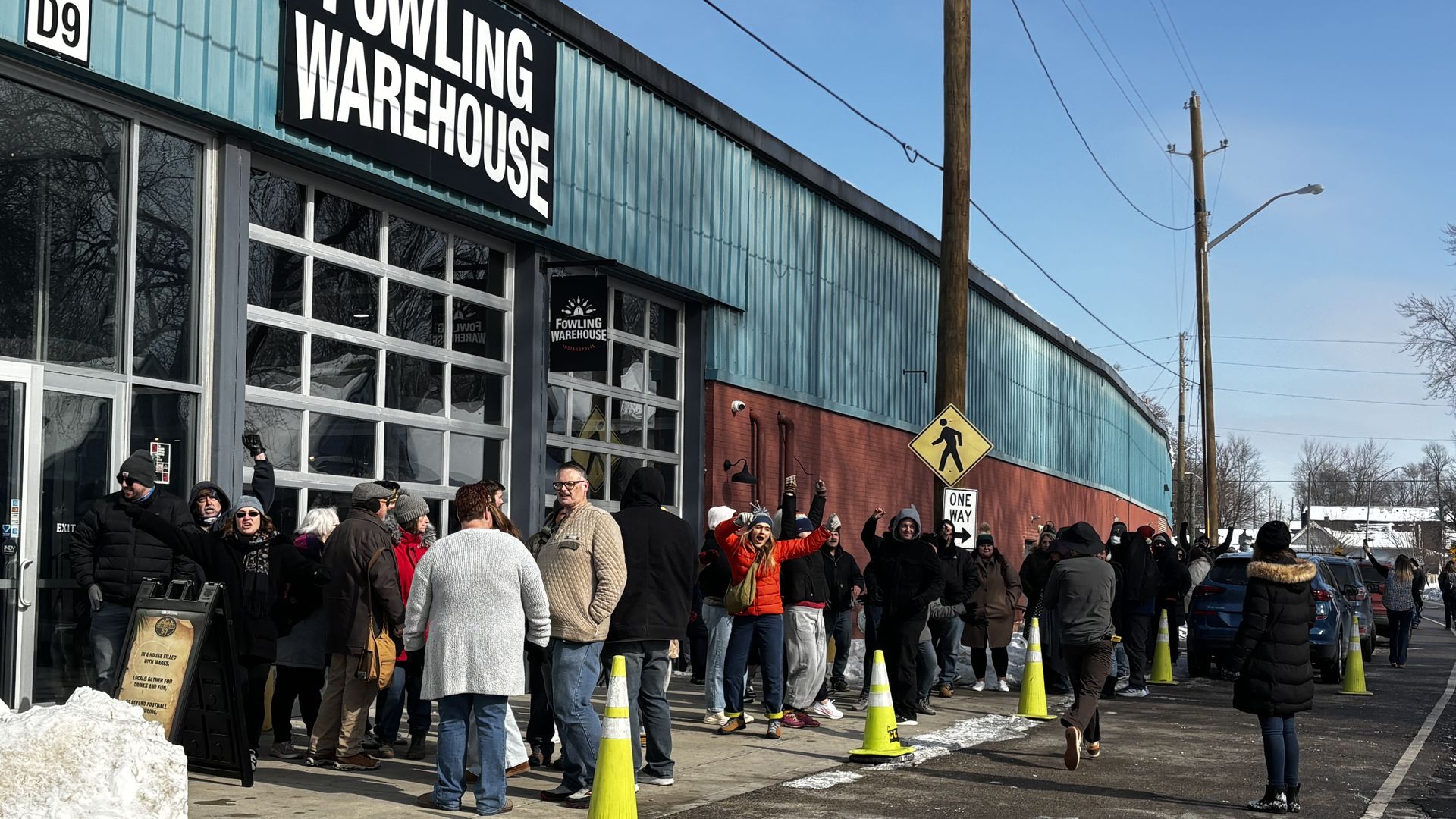People dressed in winter clothing standing in line outside a blue and red building labeled Fowling Warehouse on a clear day with snow piled nearby and yellow traffic cones along the sidewalk.