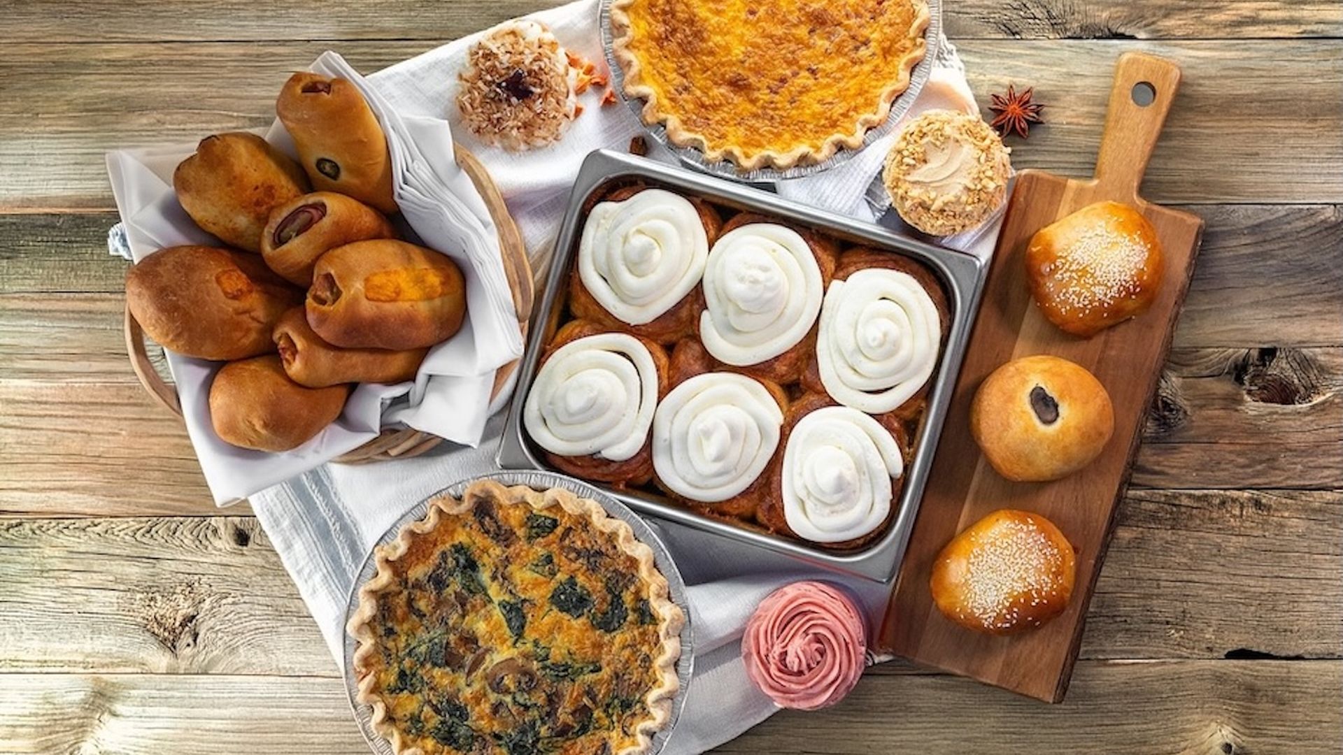 Assorted baked goods on a rustic wooden table: cinnamon rolls with white frosting, two quiches, a pie, a basket of dinner rolls, buns on a board, and a pink frosted cupcake.