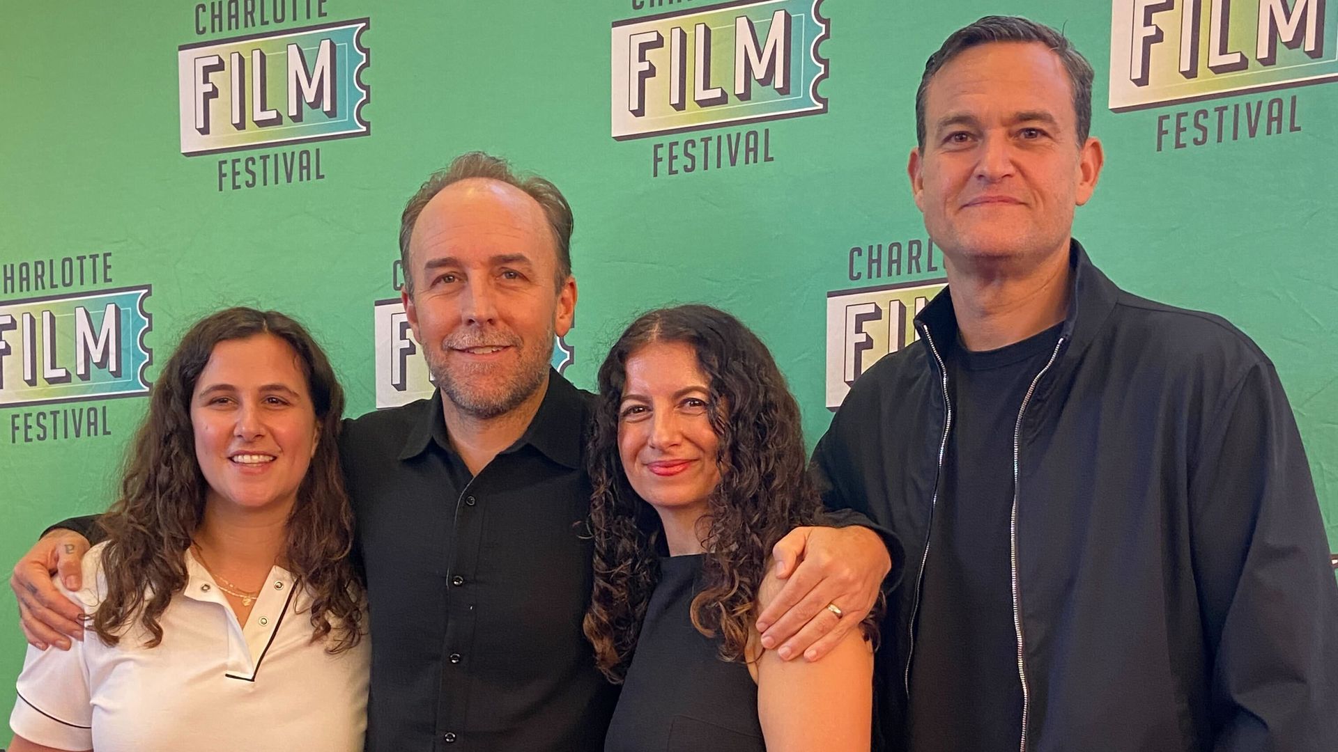 Four people standing close together on a red carpet in front of a green "Charlotte Film Festival" backdrop. Two women and two men smiling; one woman holds a shiny gold clutch.