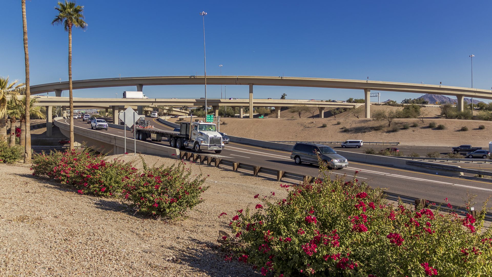 Wide desert freeway with elevated beige ramps and light poles. A white semi-truck and cars travel below; palm trees on the left, pink bushes in the foreground, blue sky and distant mountains.
