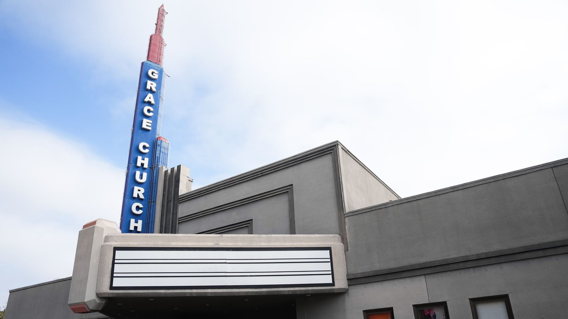 Exterior of a gray concrete building with a tall blue vertical sign reading "GRACE CHURCH". A white marquee extends over the entrance; a cloudy pale-blue sky fills the background.