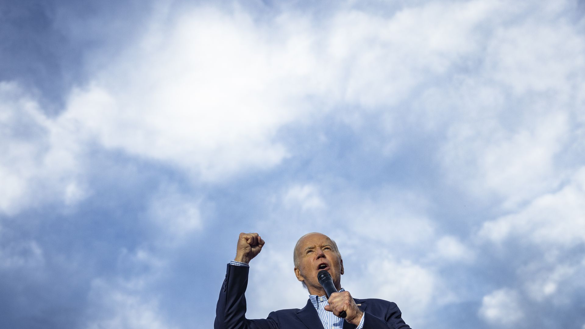 Joe Biden speaks into a microphone with a partly cloudy sky behind him.