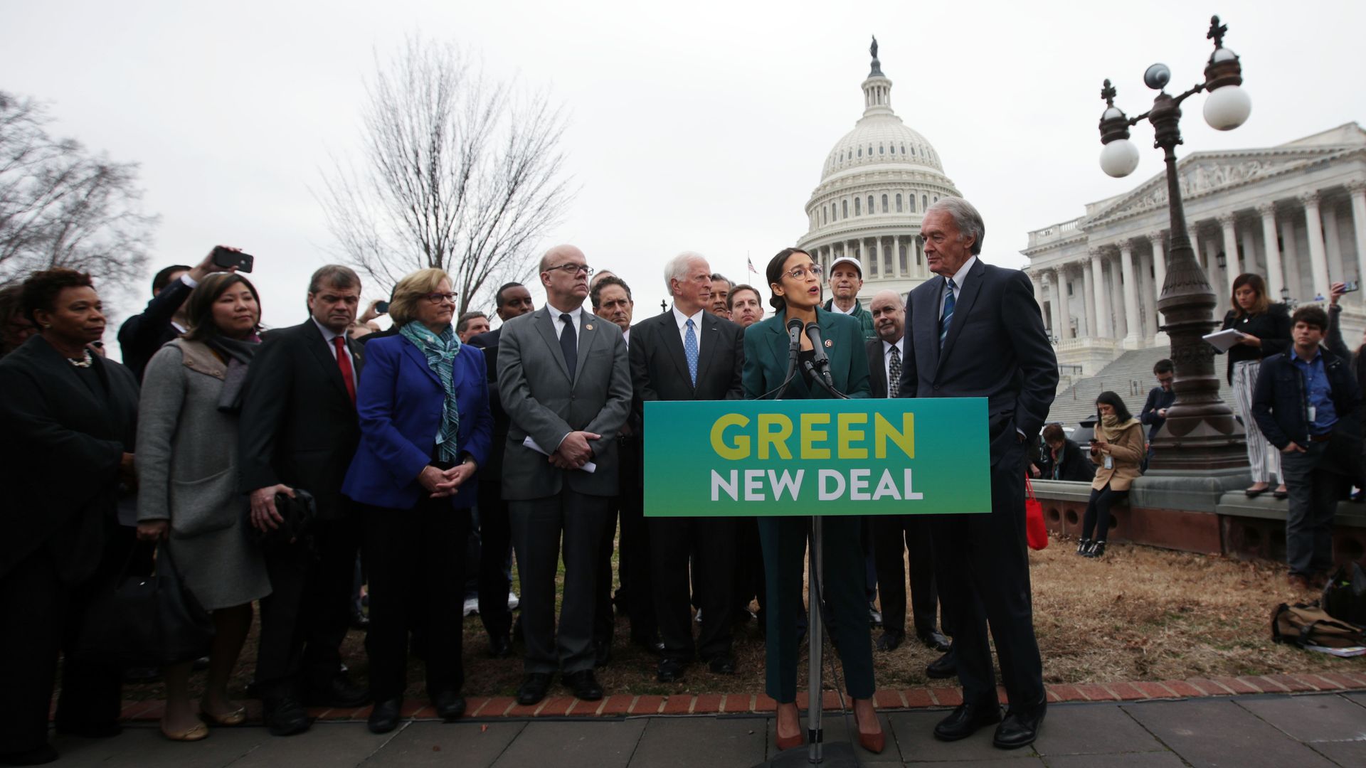 In this image, Alexandria Ocasio-Cortez stands alongside other Democrats outside the Capitol building on a sunny day in Washington. They hold a banner that says "Green New Deal." 