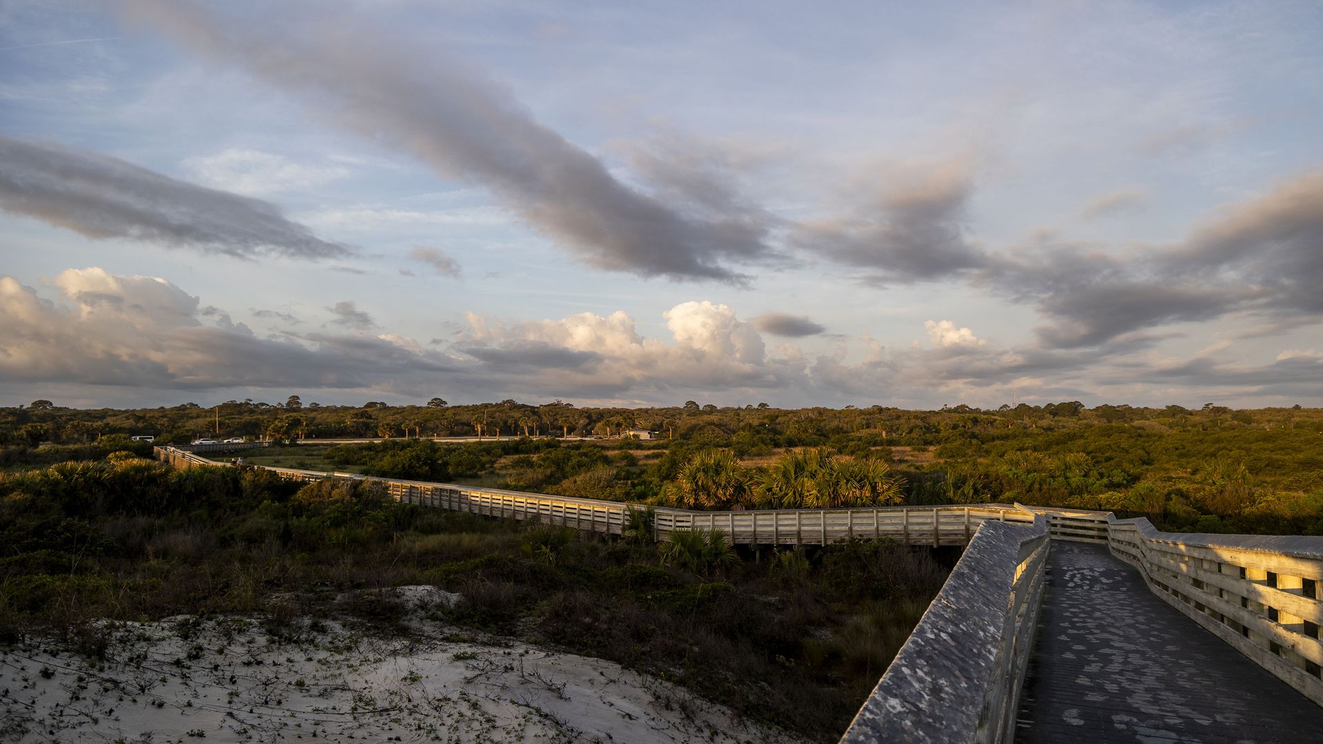A boardwalk leads over sand dunes toward a parking lot in the distance, surrounded by shrubs and trees.