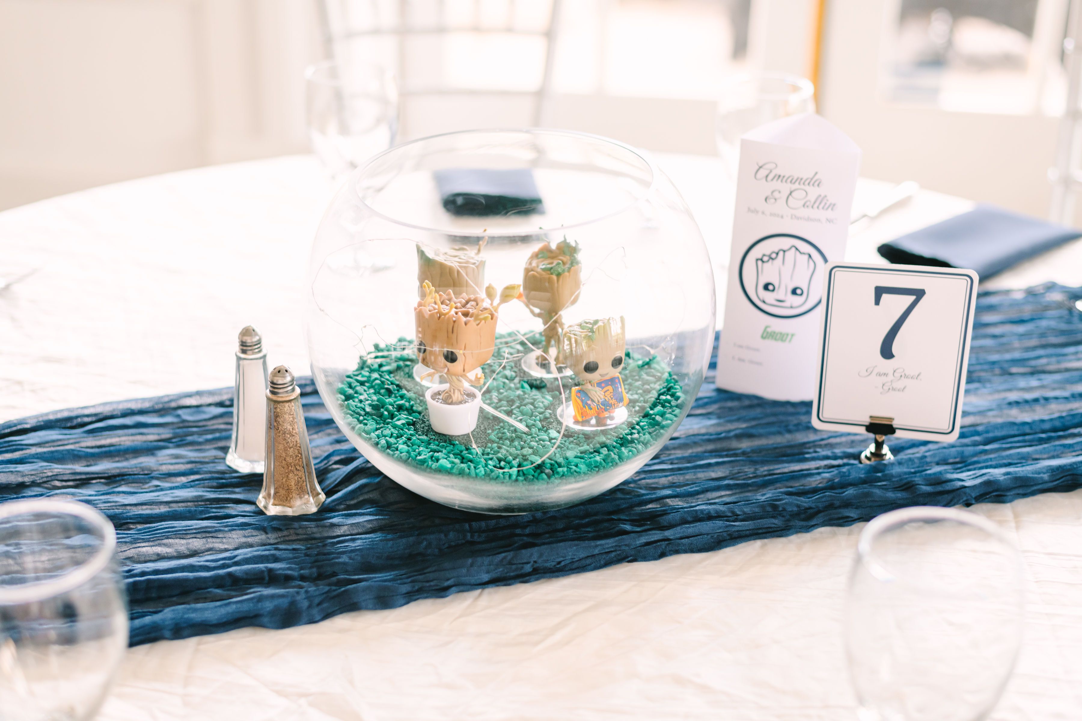 Wedding table decoration with a clear bowl containing four Groot figurines on teal pebbles, a salt and pepper shaker, a blue cloth runner, and a table card marked 7 with "I am Groot" text.