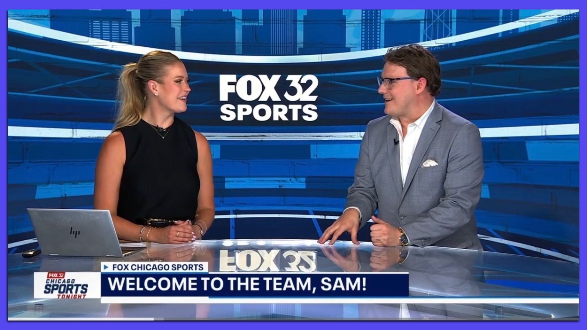 Two anchors sit at a glass desk on a blue Fox 32 Sports set; woman in a black sleeveless top chats with a man in a gray suit and glasses, laptop visible, ticker reads "Welcome to the Team, Sam!"
