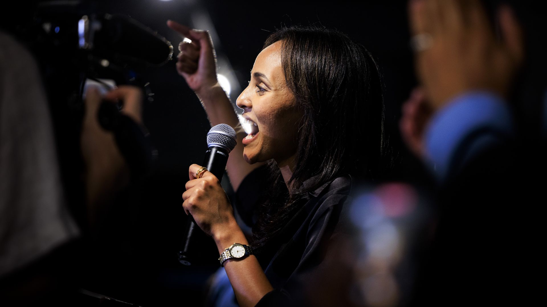 Profile view of a woman speaking into microphone. Dark background with bright spotlights and raised hands in the crowd; she wears a gold watch and rings.