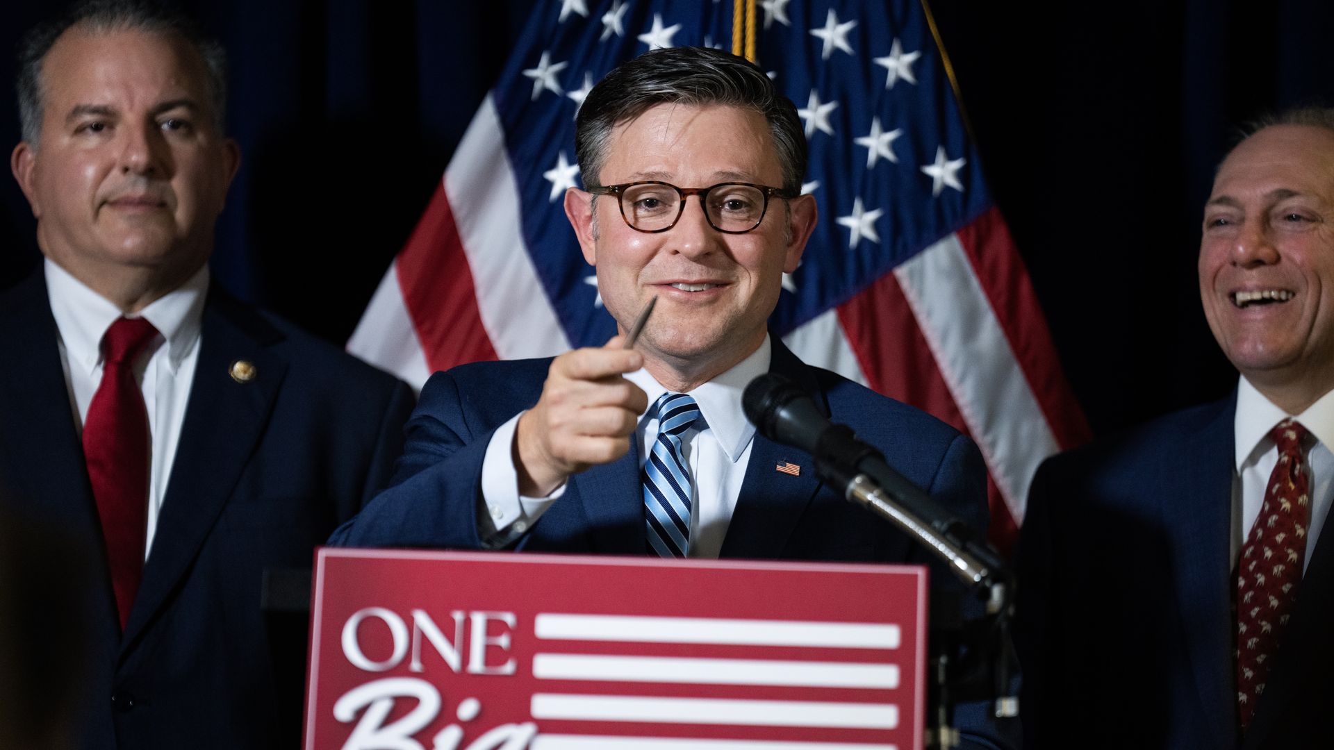 House Speaker Mike Johnson speaking at a podium with a red sign while holding up a pen, flanked by GOP colleagues with an American flag behind him.