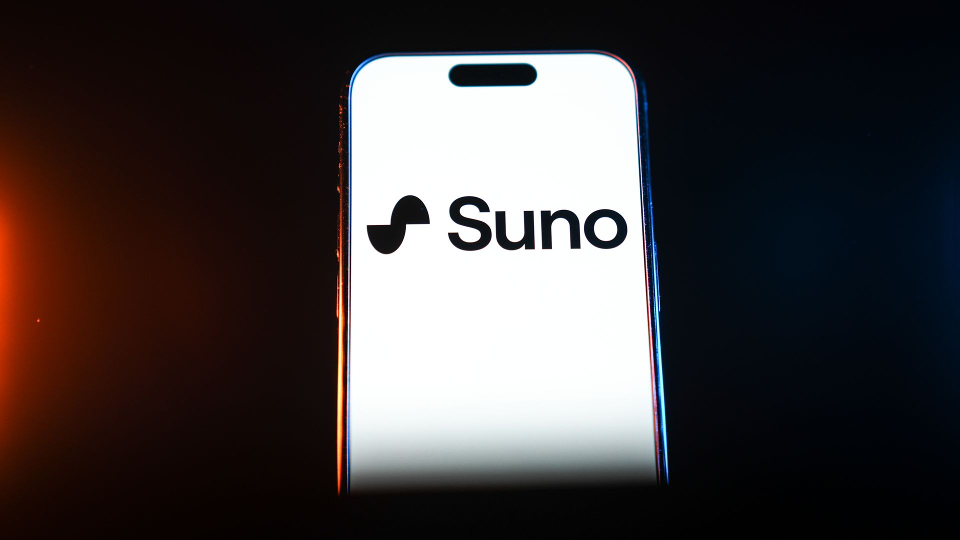 Close-up of a smartphone with a white screen showing the logo and text Suno with a rounded black symbol to the left, lit by warm orange on the left and cool blue on the right against a dark background