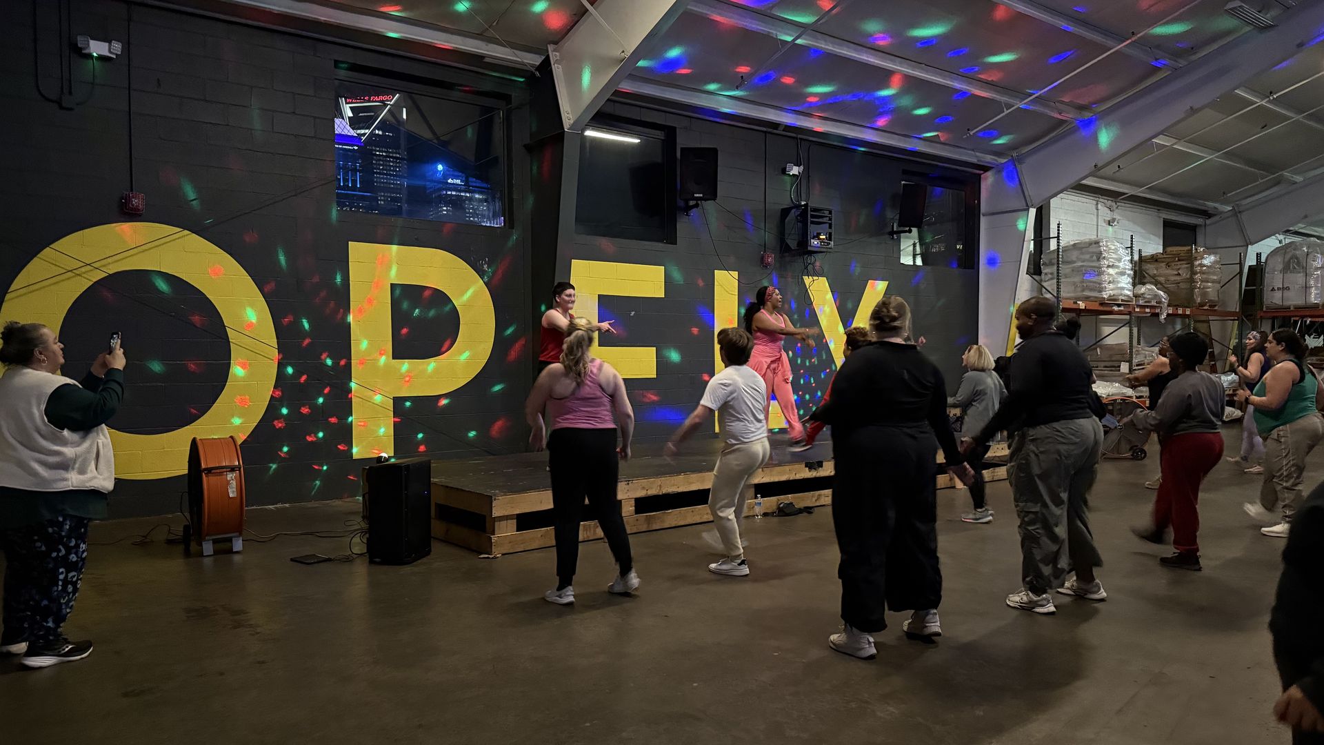 Indoor dance class with instructor and participants under colorful disco lights, next to a wall painted with large yellow letters spelling "OPENLY" in a warehouse-like space.