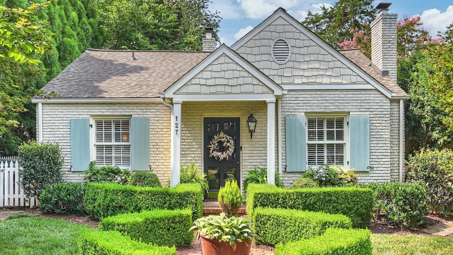 Hot homes. Charlotte houses for sale. A charming single-story brick cottage with light gray shingles on the gable, blue window shutters, a black front door with a wreath, and manicured boxwood hedges along a brick path to the entrance.