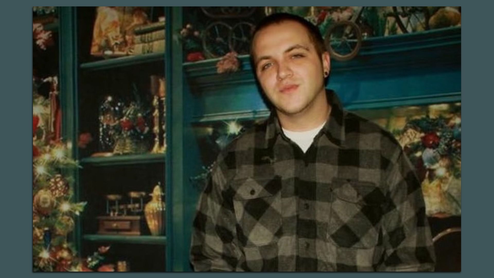 Young man in a black and gray checkered shirt stands indoors near a decorated Christmas tree and antique-style shelves with books and ornaments, with a slight smile on his face.