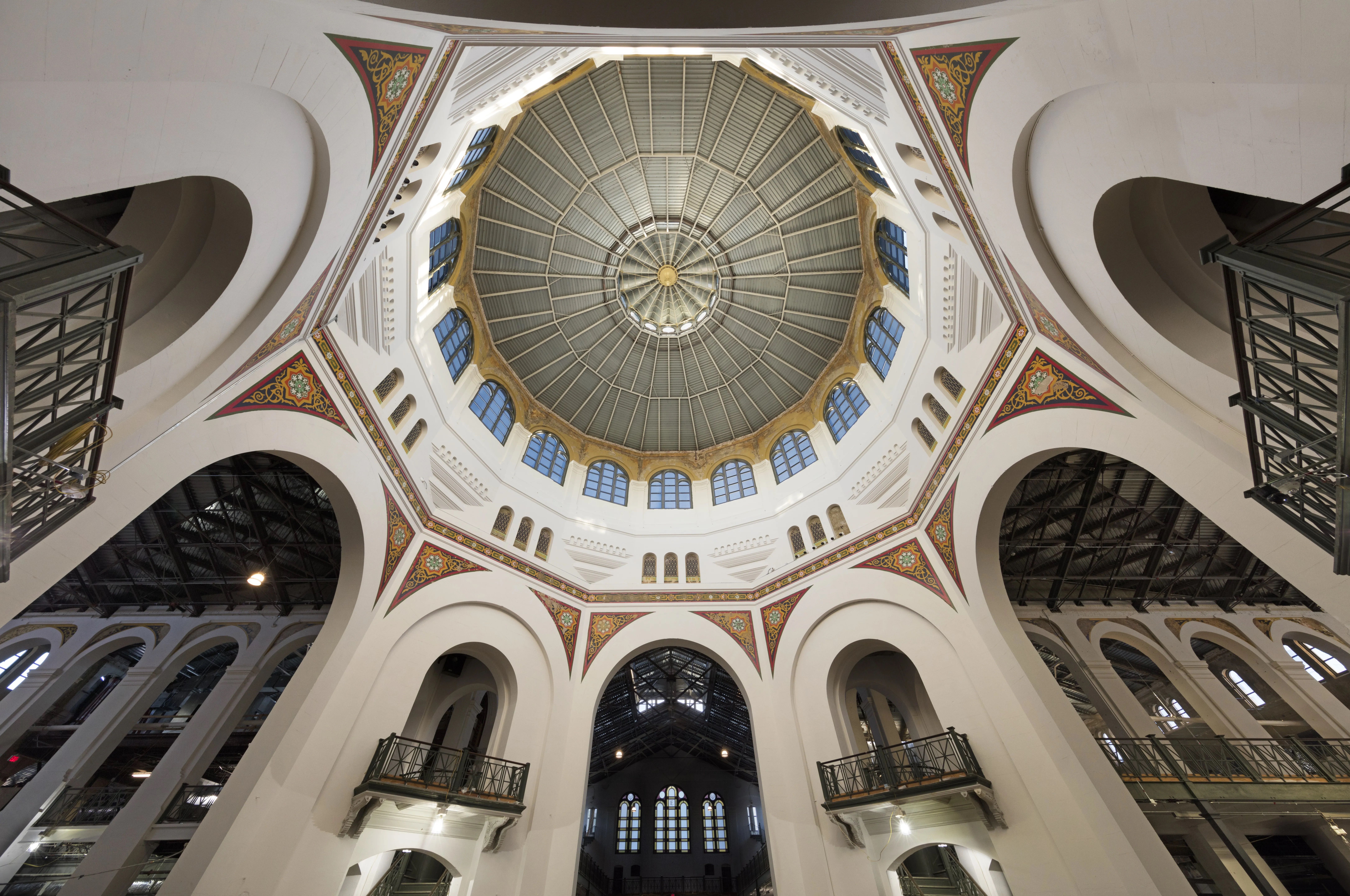 Interior of a grand domed hall viewed from below: a circular, segmented glass dome with a gold center, flanked by white arches, intricate red-gold decorative panels, and metal balconies.