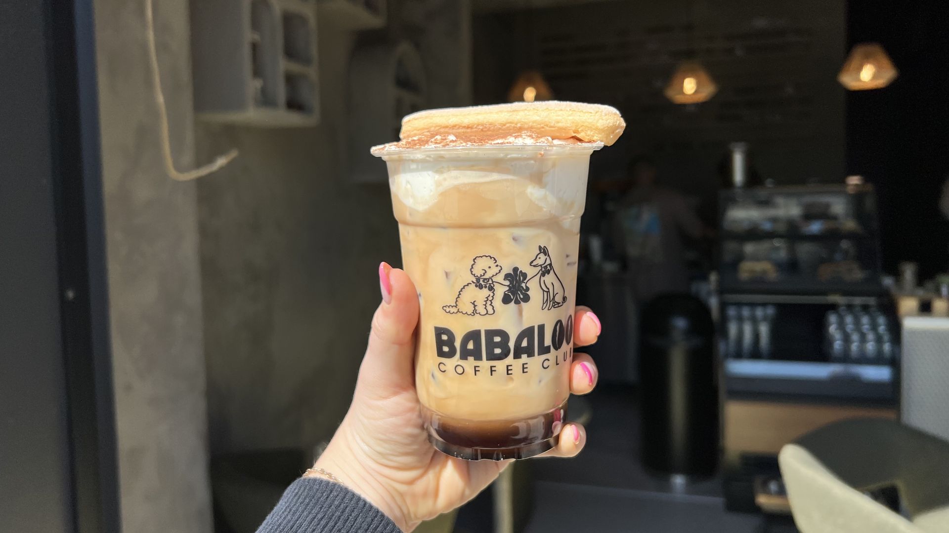 Hand with pink nails holds a plastic cup of iced coffee topped with a round biscuit; the cup shows cartoon animals. A cozy cafe interior with warm pendant lights is blurred in the background.