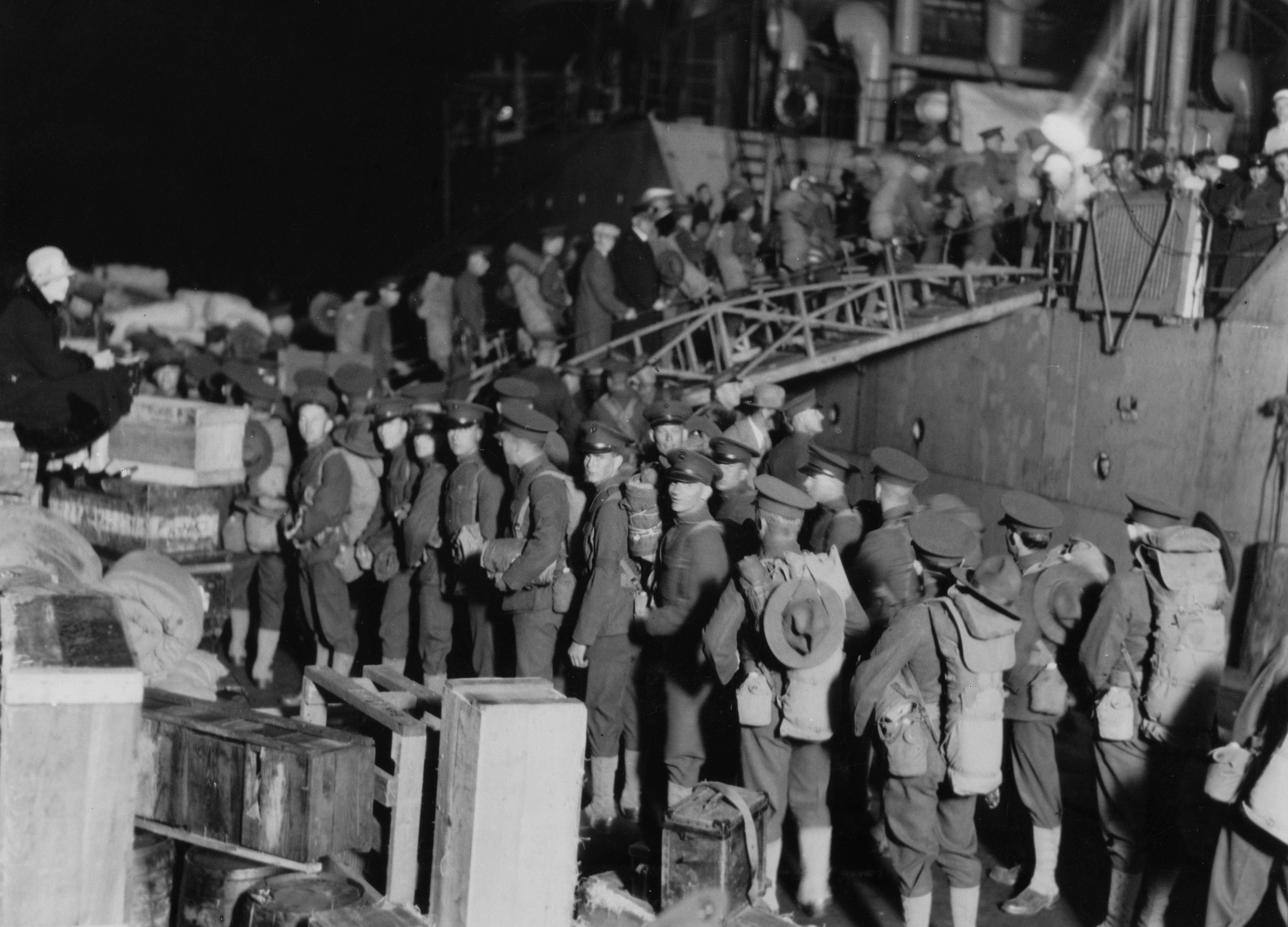 Black-and-white photo of a long line of soldiers in uniform with peaked caps standing along a wooden barrier beside a large vessel. Civilians and crew watch from the deck; a man in a suit leans on a railing in the foreground.