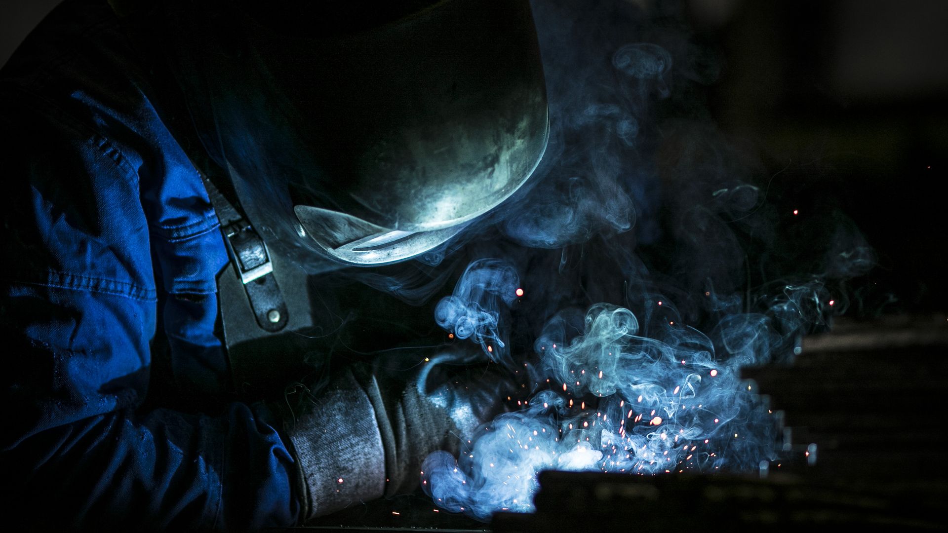 A worker melds medal in a dark room with blue light exposing some smoke.