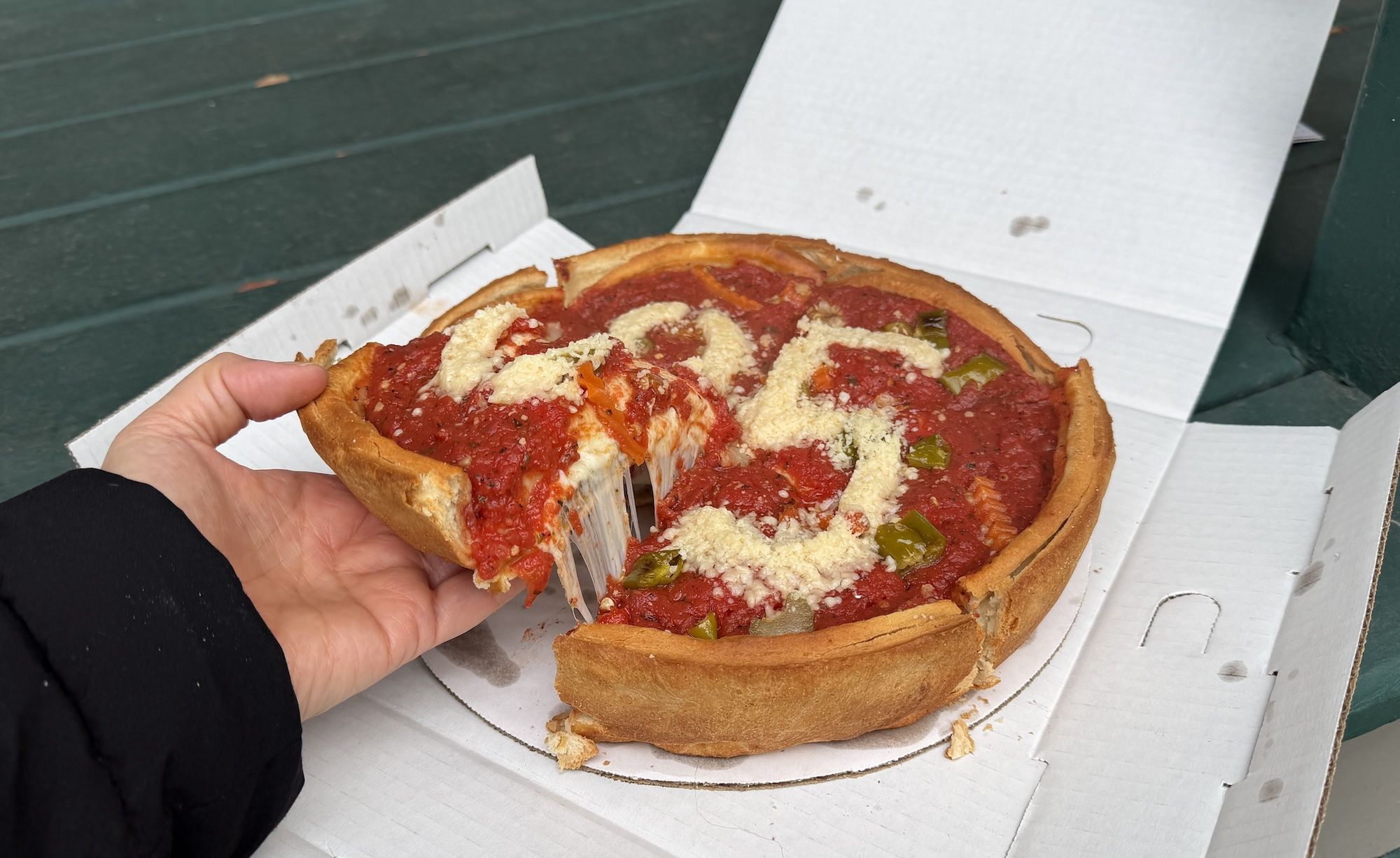 Hand holding a slice of deep-dish pizza with tomato sauce, green peppers, and grated cheese forming the numbers "425" on top in a white pizza box.