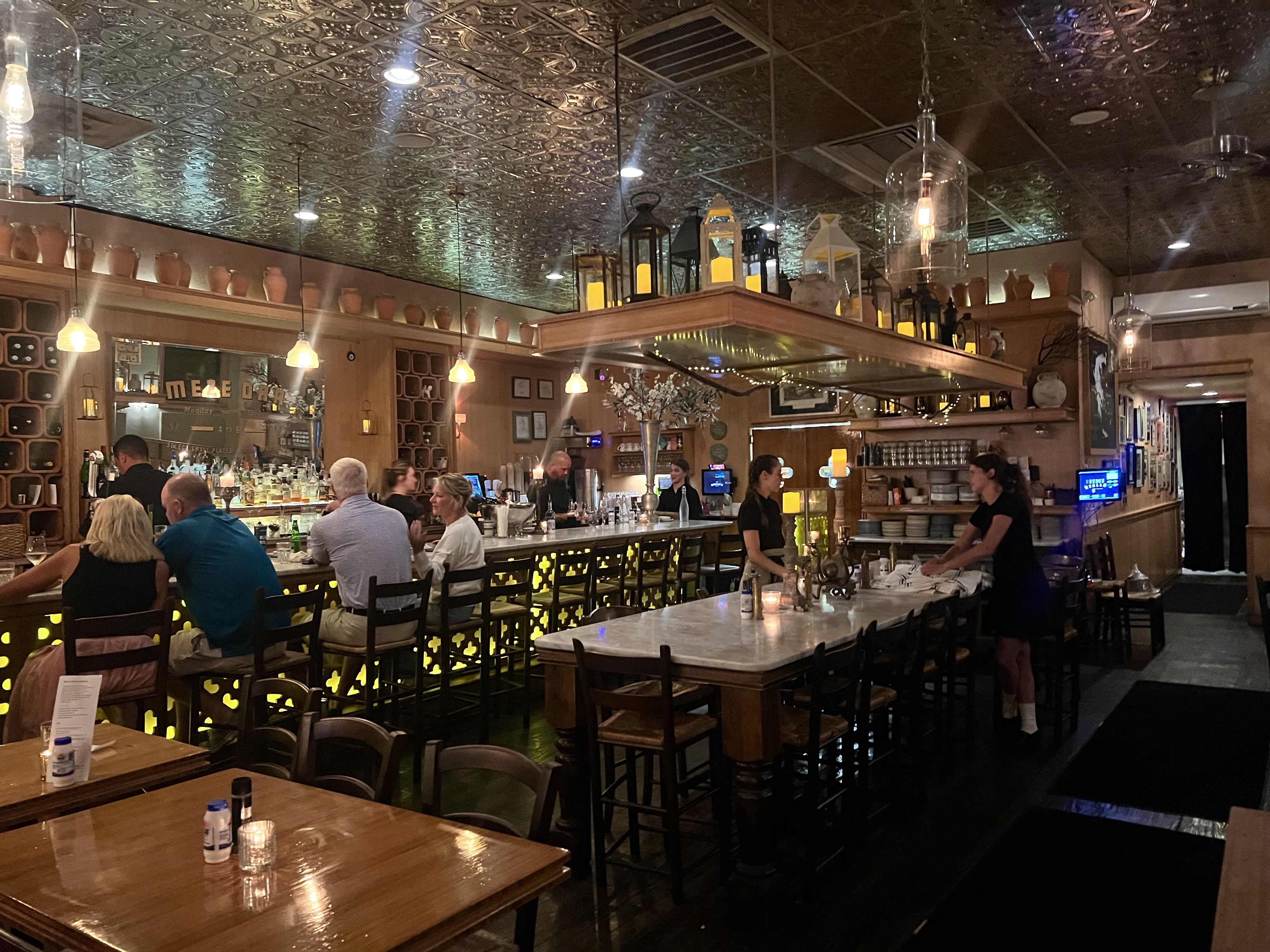Cozy bar interior with patrons seated at a lighted bar counter, ornate ceiling, warm yellow lighting, a large communal table with candles, and staff preparing around it.