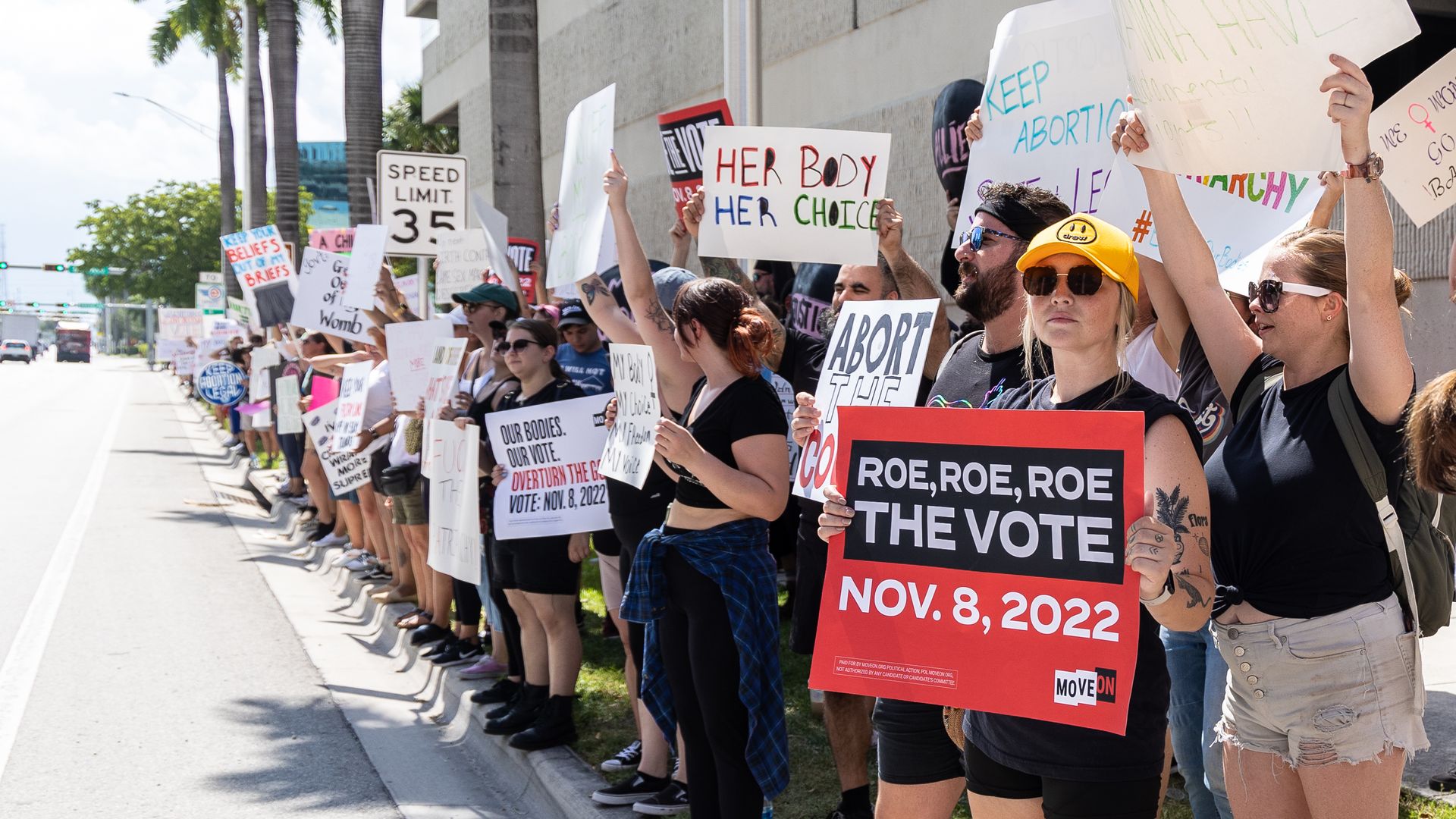 A group of protesters stand on a sidewalk holding signs. Among the messages are "Roe, Roe, Roe the vote" and "her body her choice."