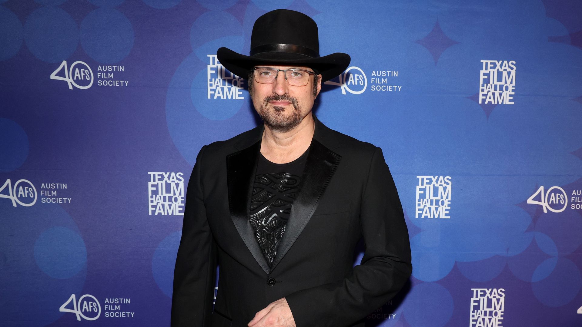 Robert Rodriguez, wearing a black jacket and cowboy hat, poses in front of a blue stop-and-repeat.