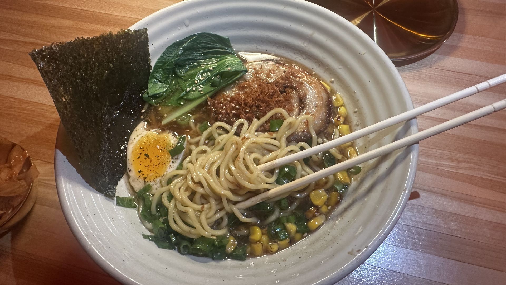 Top-down bowl of ramen with seaweed, spinach, corn, and a halved boiled egg. Chopsticks rest on the rim; wooden table and glassware visible in the background.