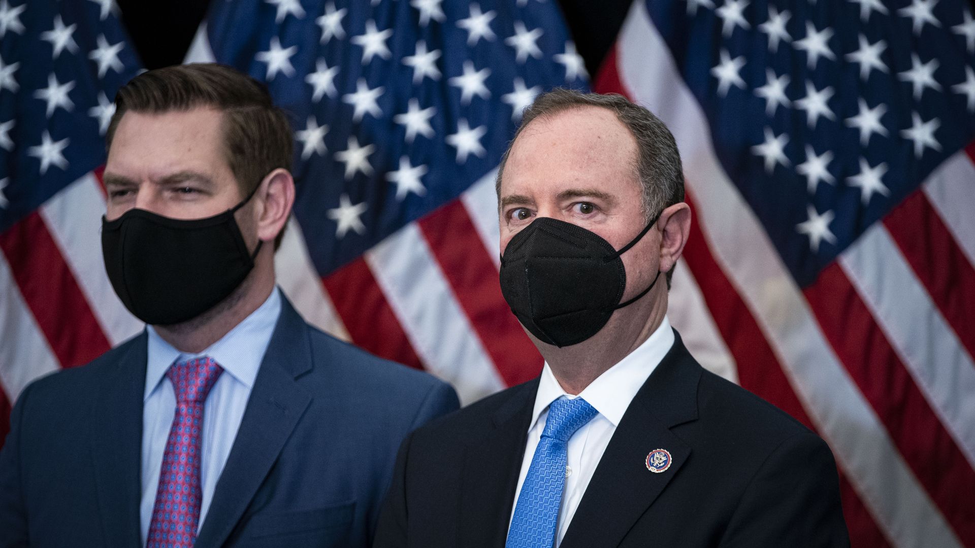 Reps. Eric Swalwell – wearing a black mask blue suit, blue shirt and purple tie – and Adam Schiff – wearing a black mask, blue suit, white shirt and blue tie – standing in front of American flags.