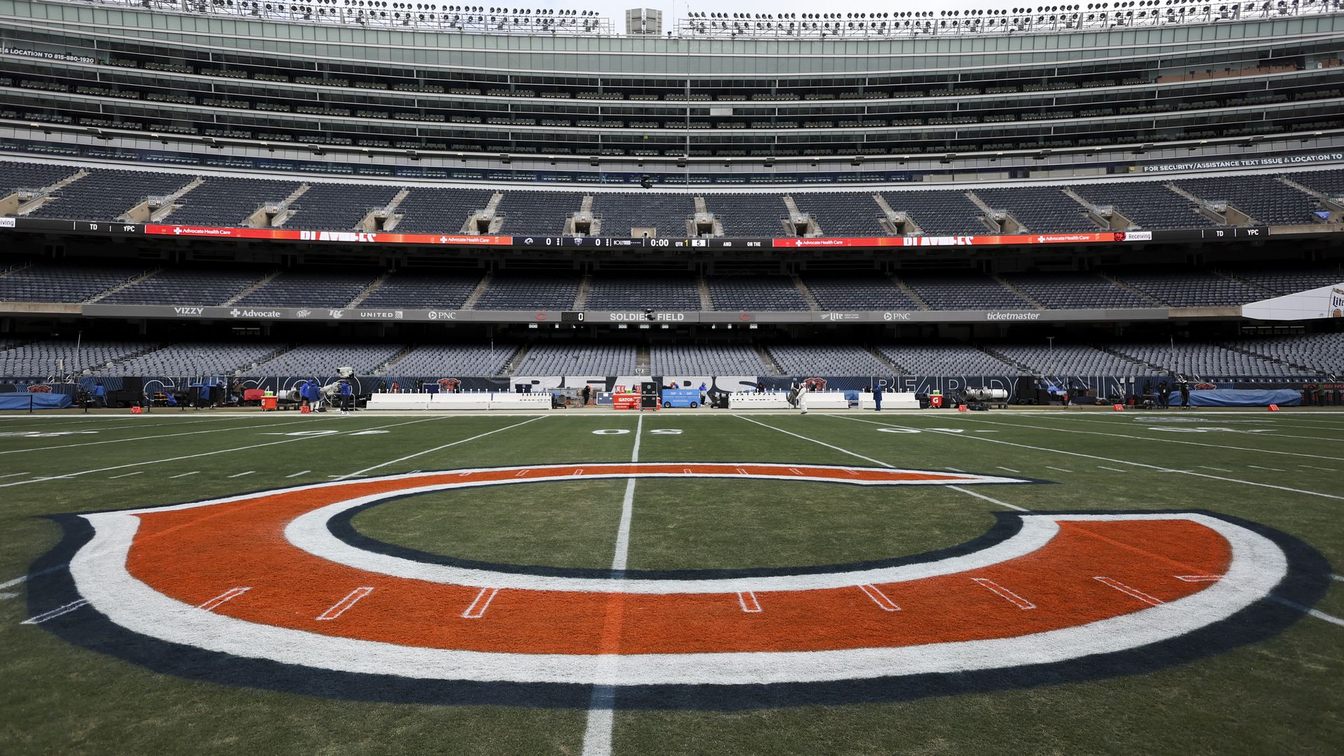 Empty Soldier Field football stadium with Chicago Bears orange and navy logo on green field under overcast sky and empty blue-gray seats.