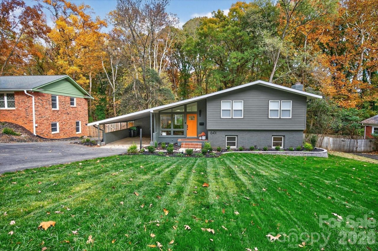 Modern gray split-level house with bright orange front door, sloped roof, green lawn, autumn trees in background, and adjacent red brick house with green trim.